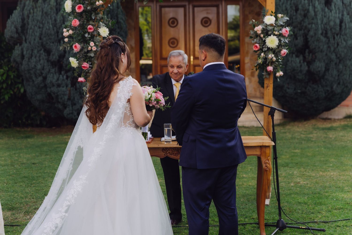 Officiant smiles as bride and groom stand together at a floral-decorated wooden arch during their wedding.