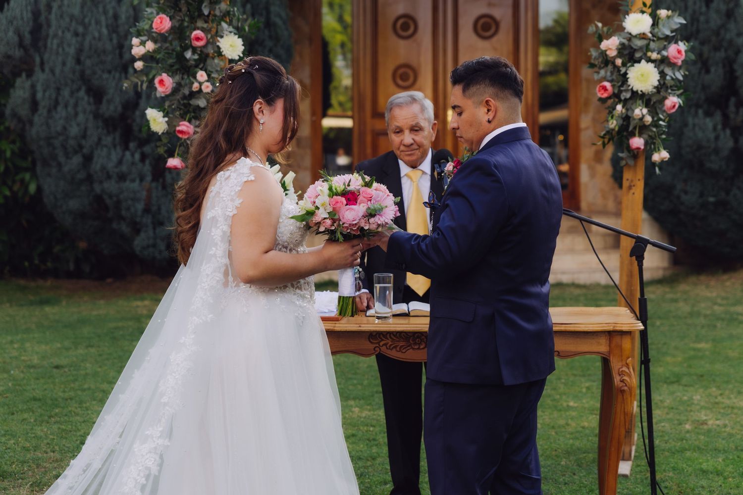 Bride and groom exchange rings with the officiant at an outdoor wedding ceremony surrounded by floral decor.