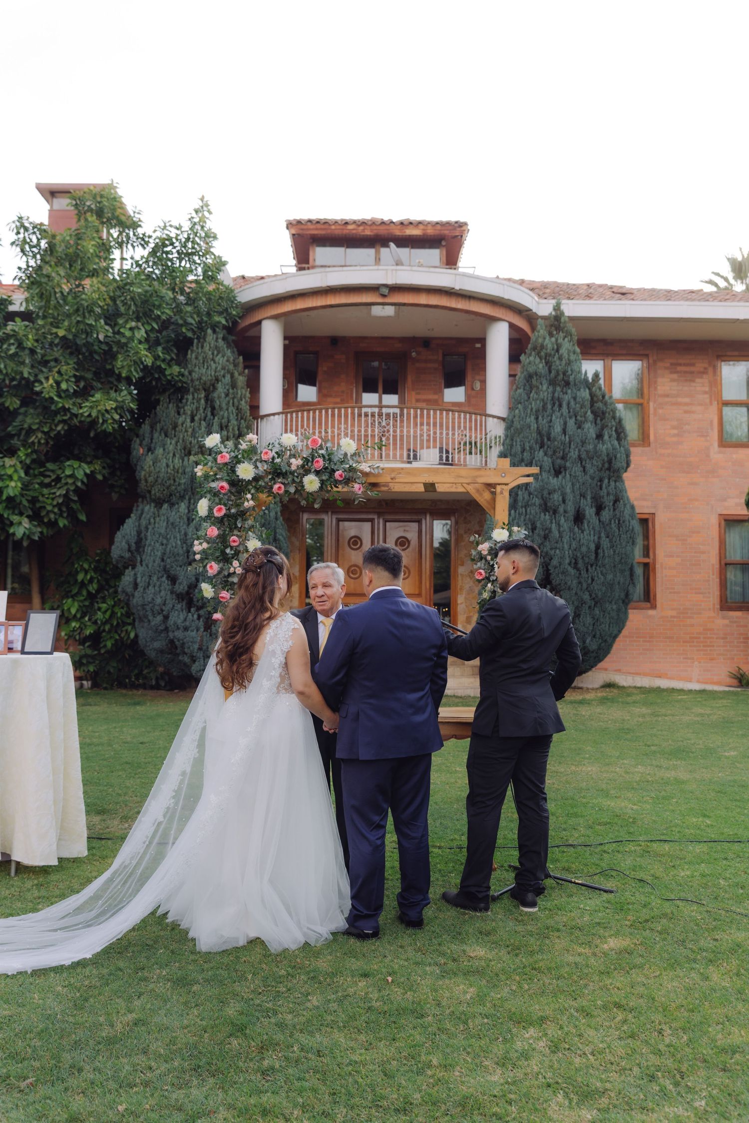 Outdoor wedding ceremony on green lawn with bride, groom, and officiant before floral arch at elegant brick mansion.