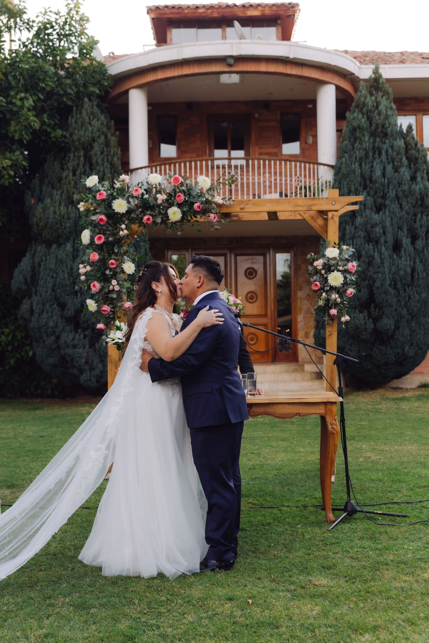 Newlyweds share their first kiss at outdoor wedding ceremony with floral arch and brick estate backdrop.