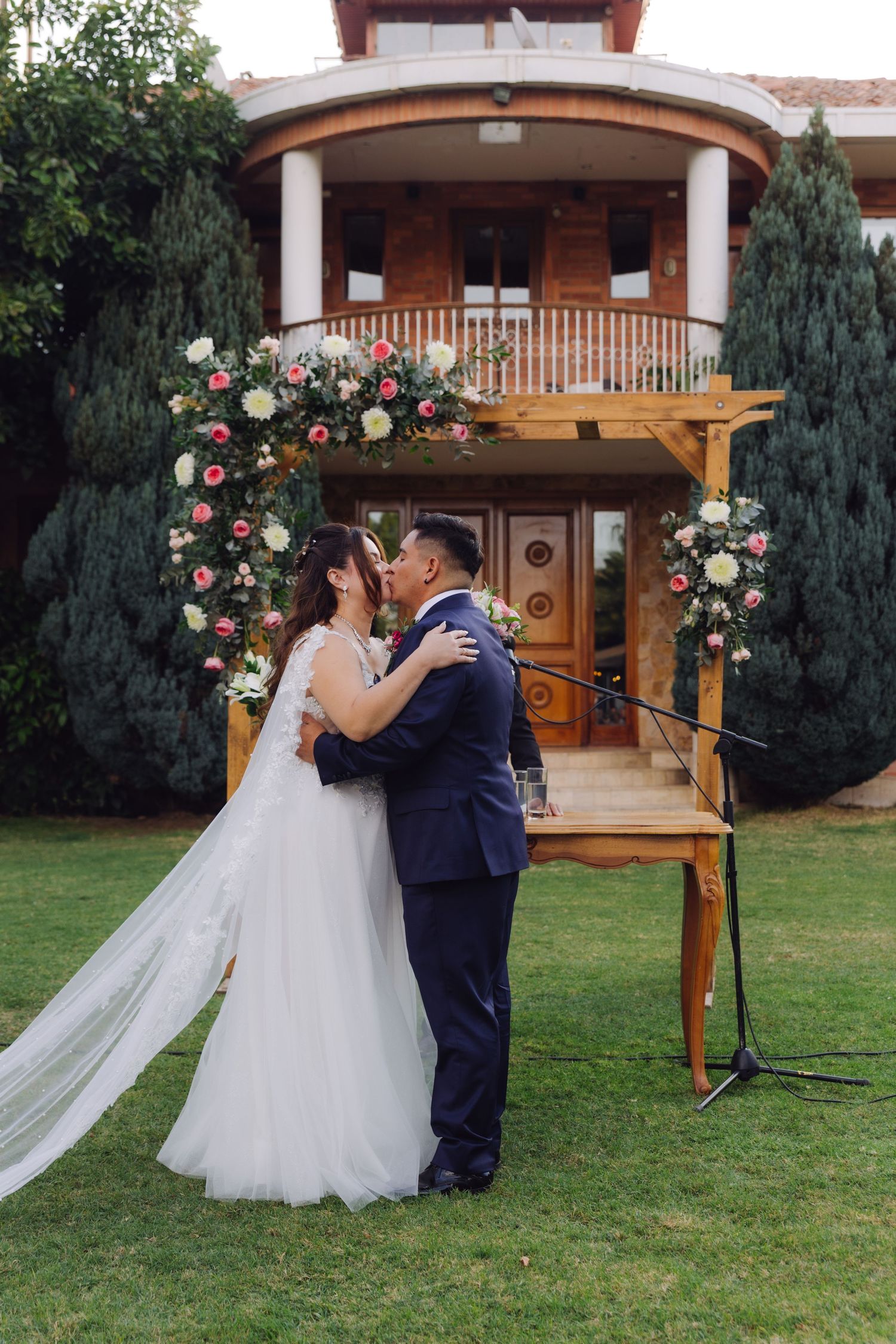 Bride and groom kissing during outdoor wedding ceremony in front of wooden floral arch at luxury venue.