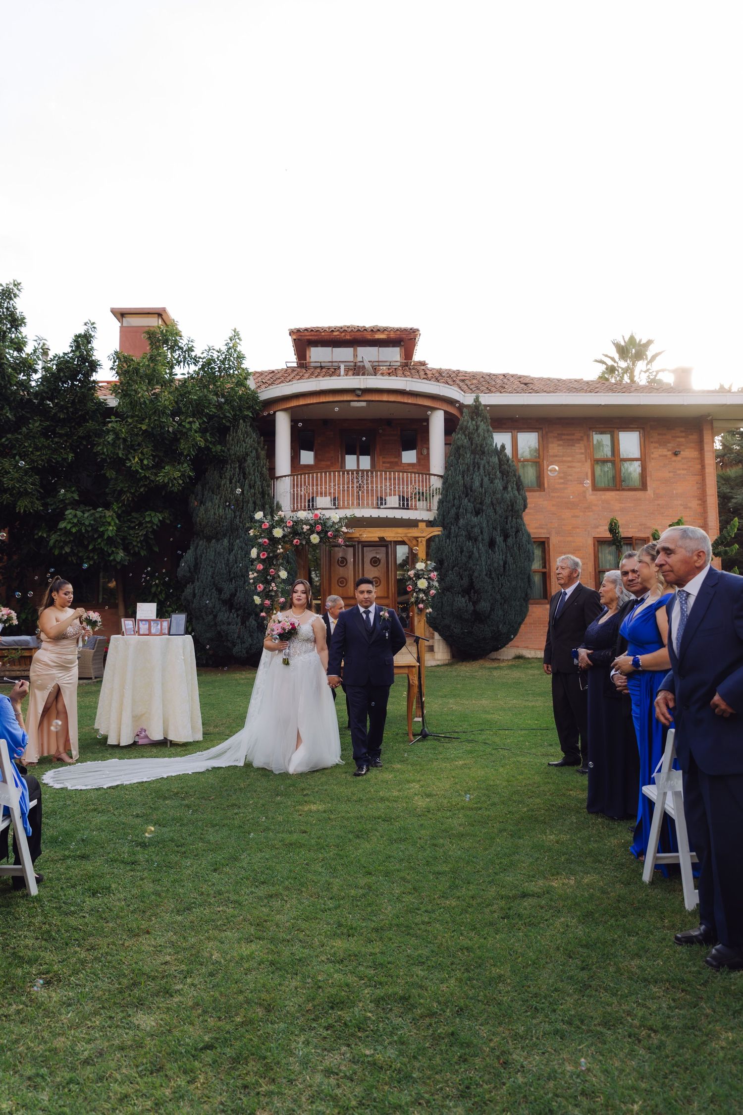 Bride walks down the aisle at sunset outdoor wedding ceremony with guests seated on white chairs.