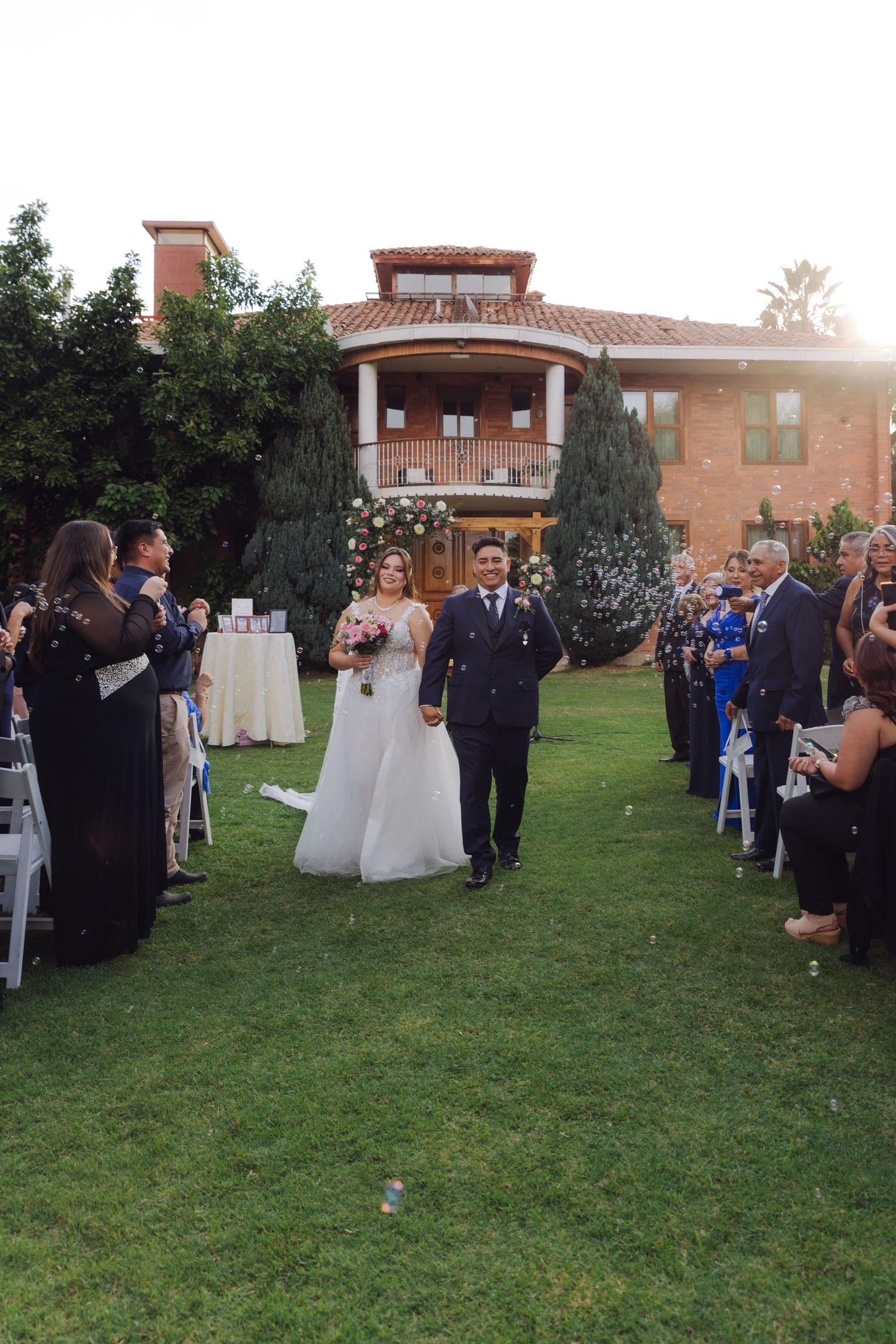 Smiling bride and groom walk down the aisle together at golden hour outdoor wedding with guests cheering.