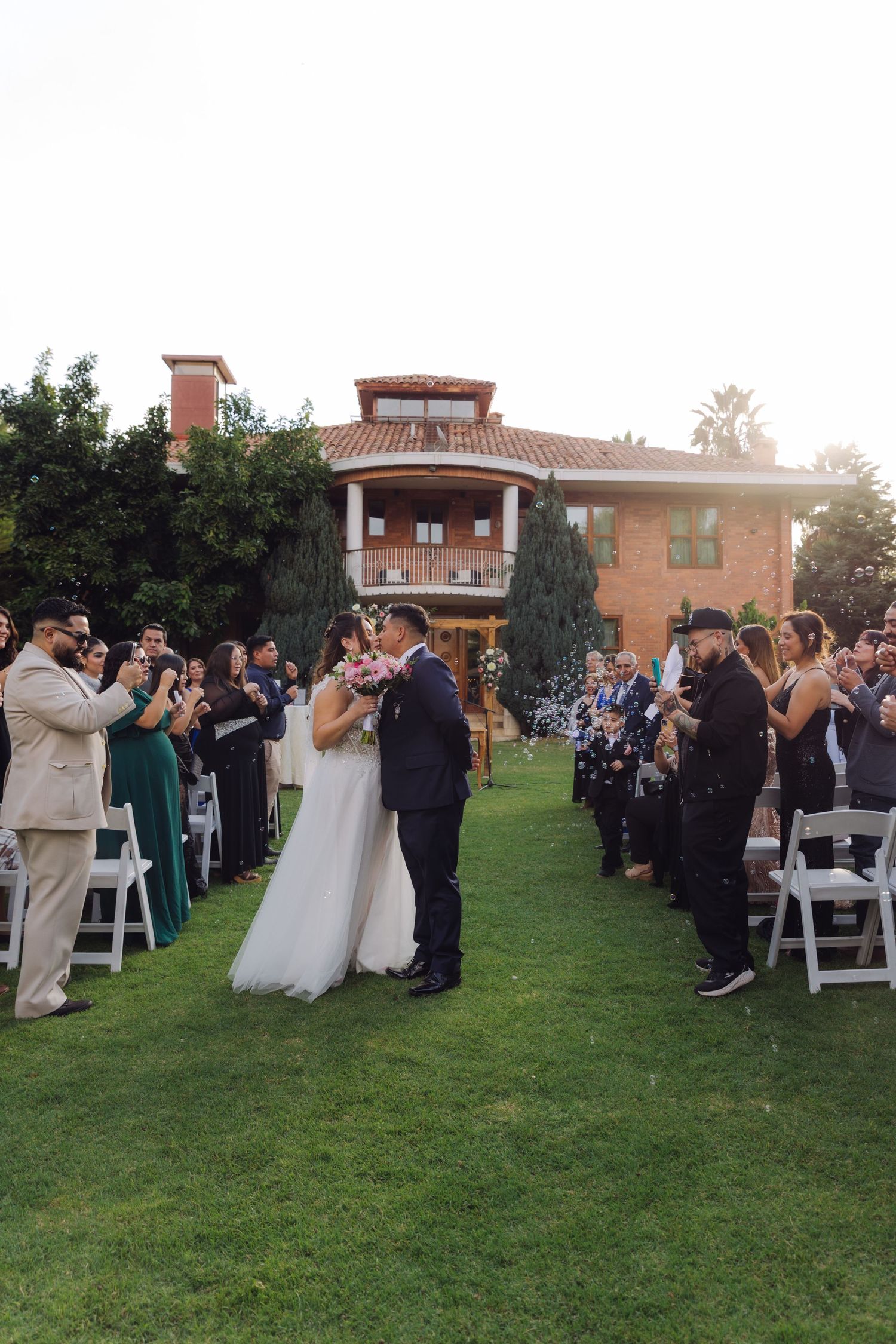 Bride and groom share a kiss while guests blow bubbles during outdoor wedding recessional at sunset.