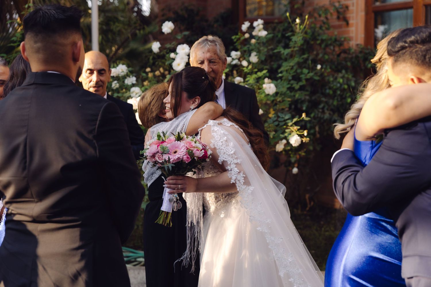 Bride receives warm congratulatory hug from guest while holding pink bouquet after outdoor wedding ceremony.