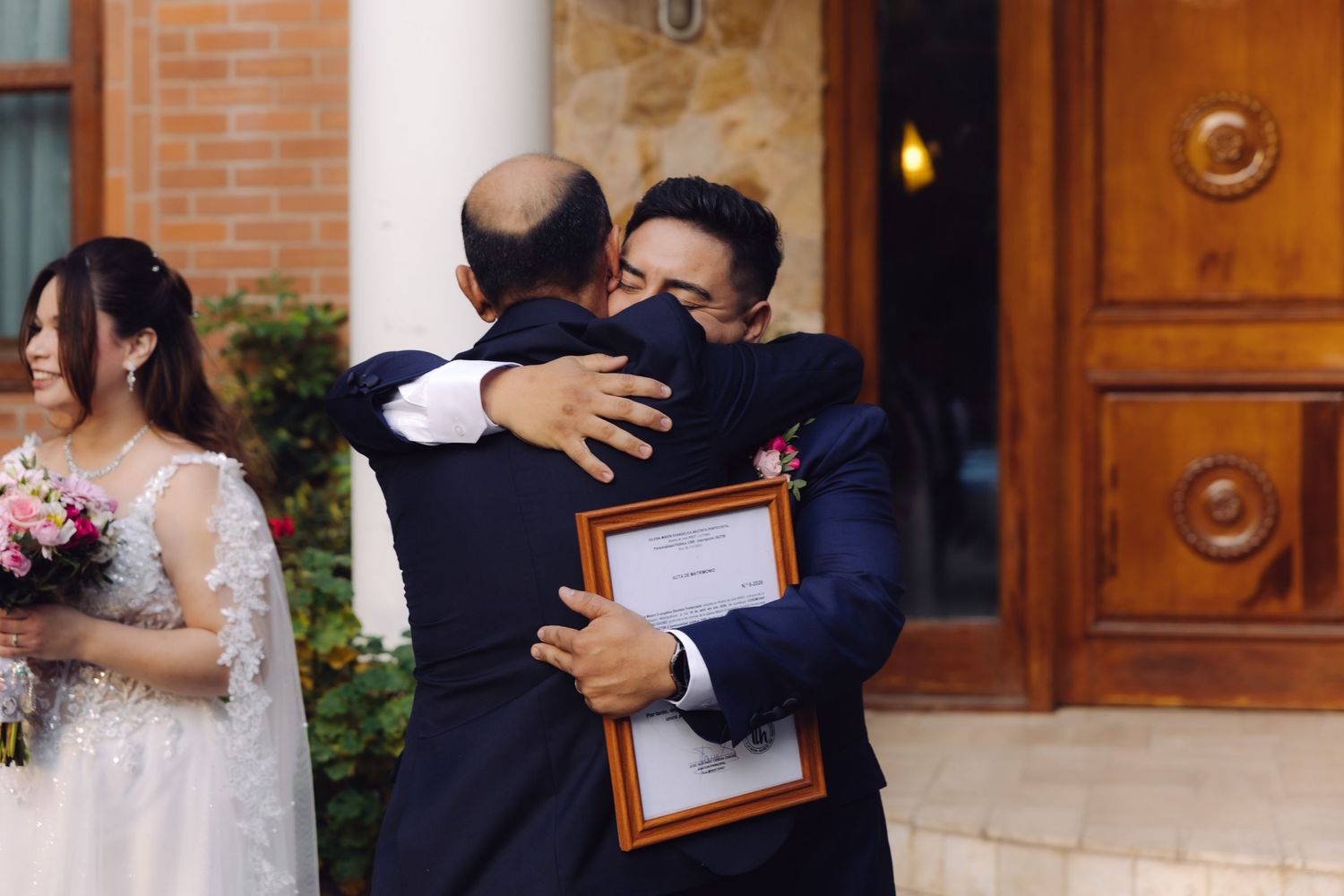 Groom hugging a guest while holding a framed wedding certificate outside a venue.