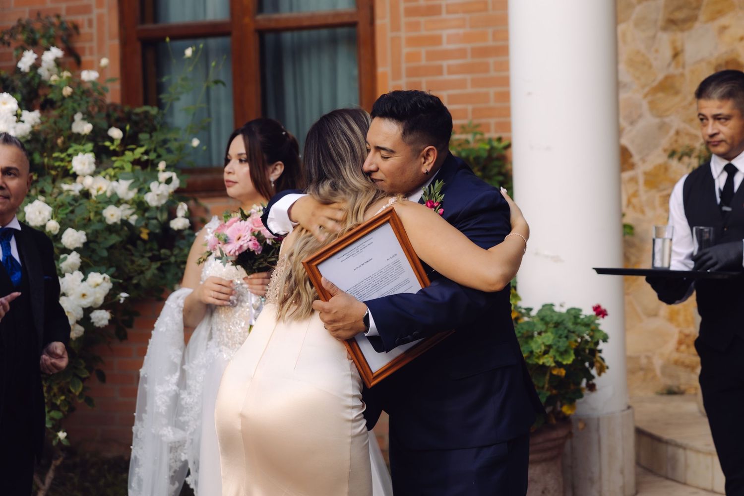 Bride hugging a guest holding a framed certificate at an outdoor wedding ceremony.