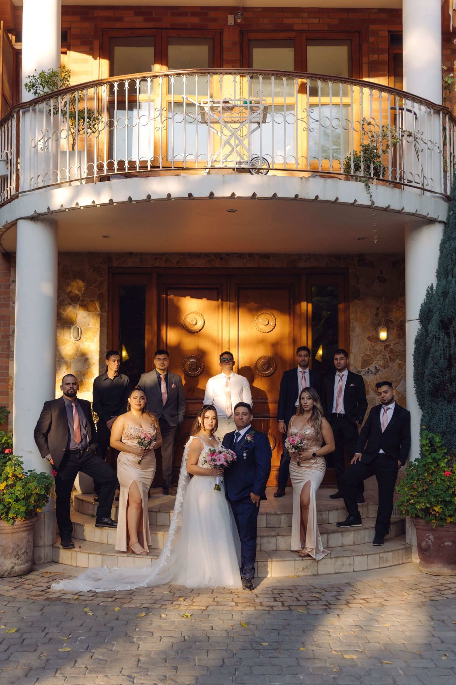 Wedding party group photo on steps of an elegant stone building at golden hour.