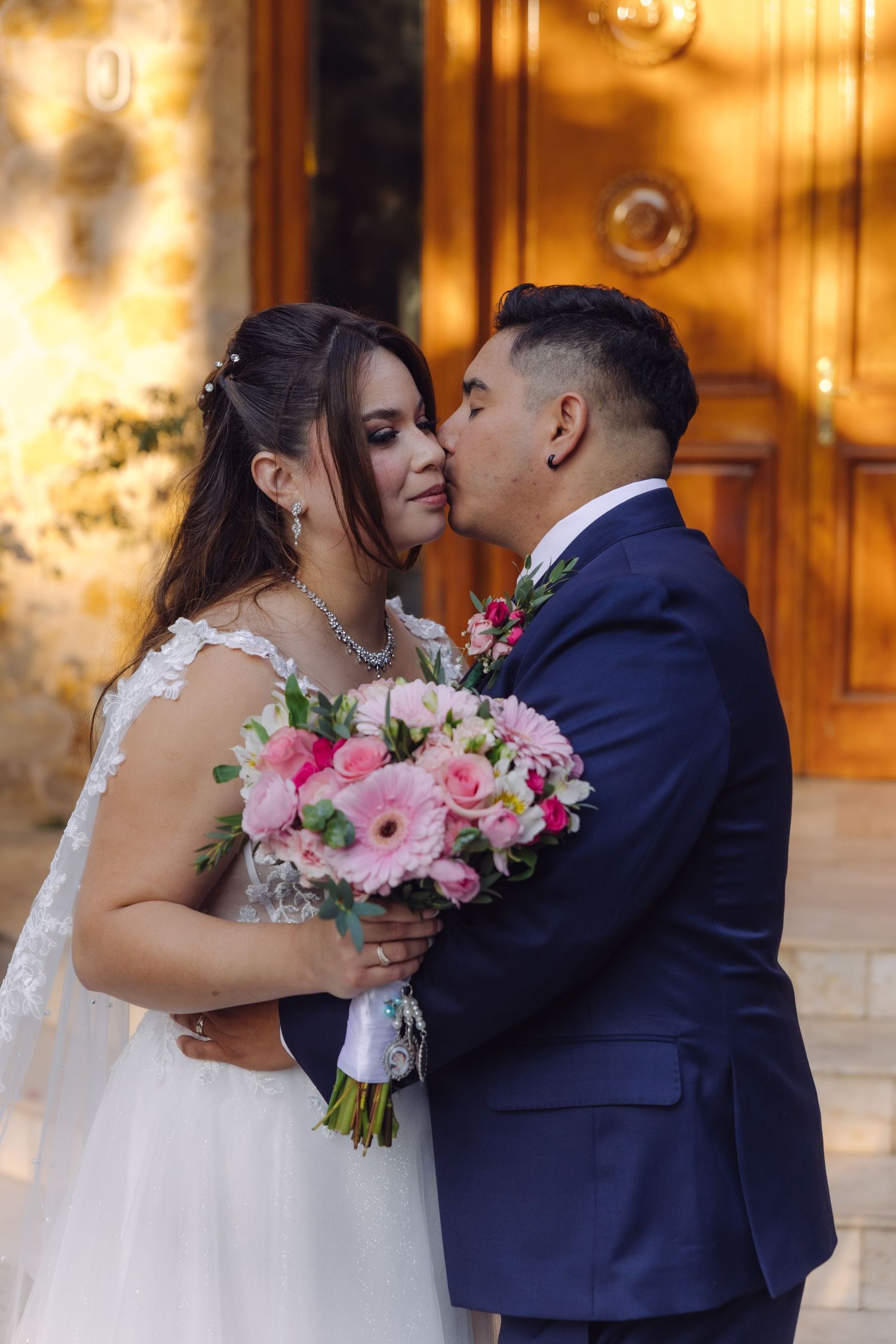 Groom kissing bride on the cheek while she holds a pink bridal bouquet.