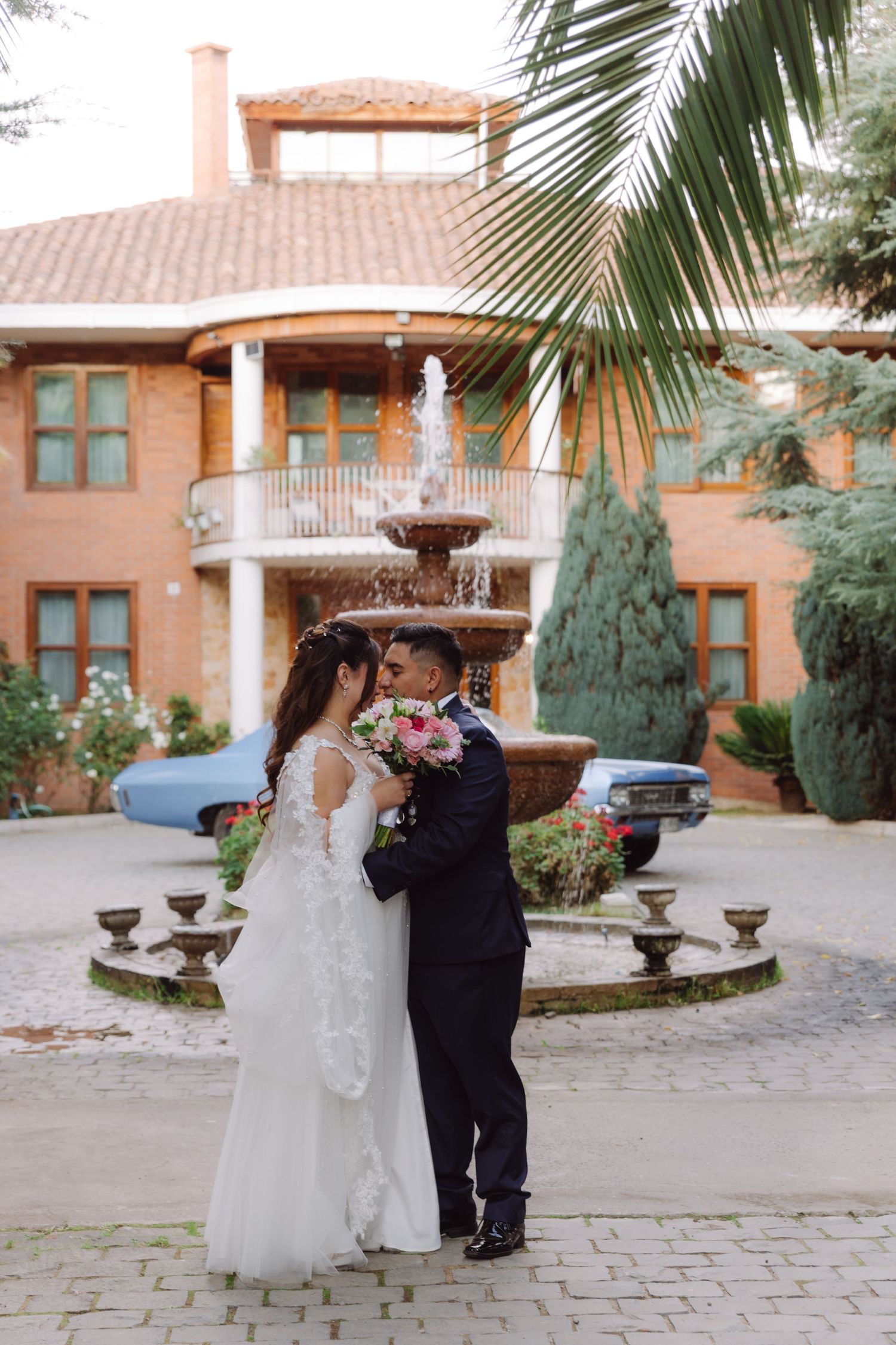 Bride and groom kissing near a fountain outside an elegant brick venue with a palm tree.