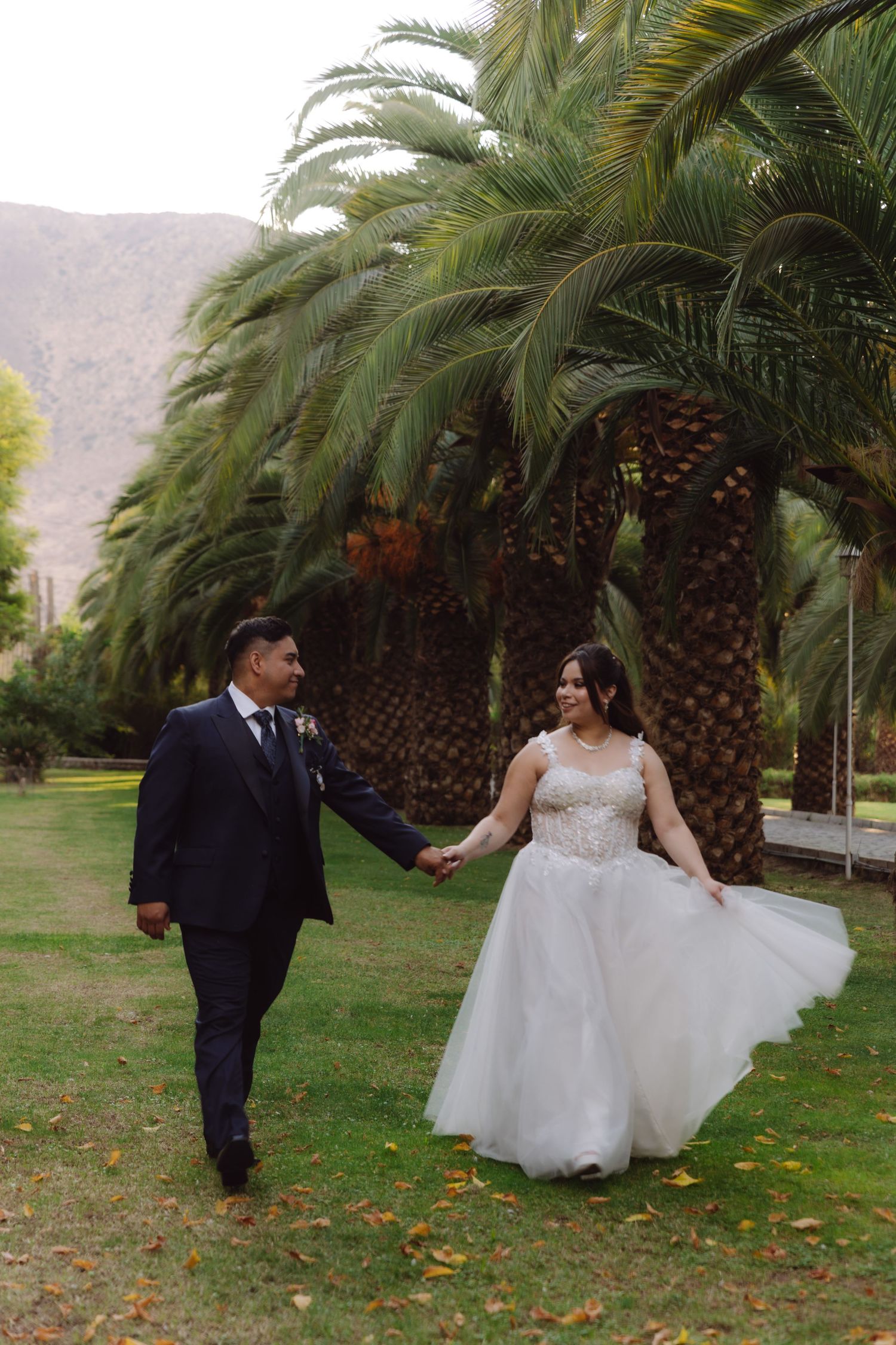 Bride and groom holding hands walking through a lush green park lined with tall palm trees.