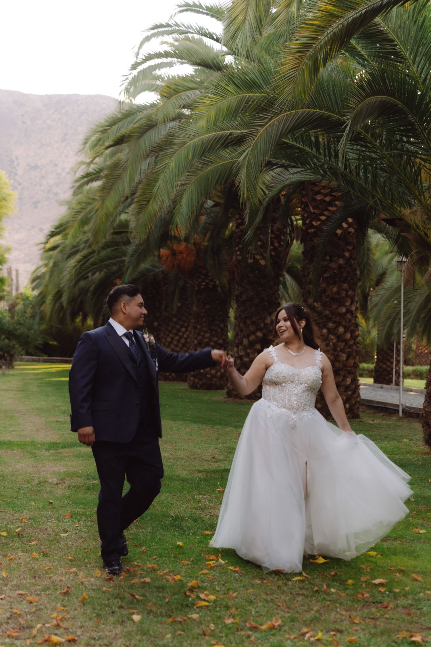 Happy couple in wedding attire holding hands and smiling while strolling under palm trees.