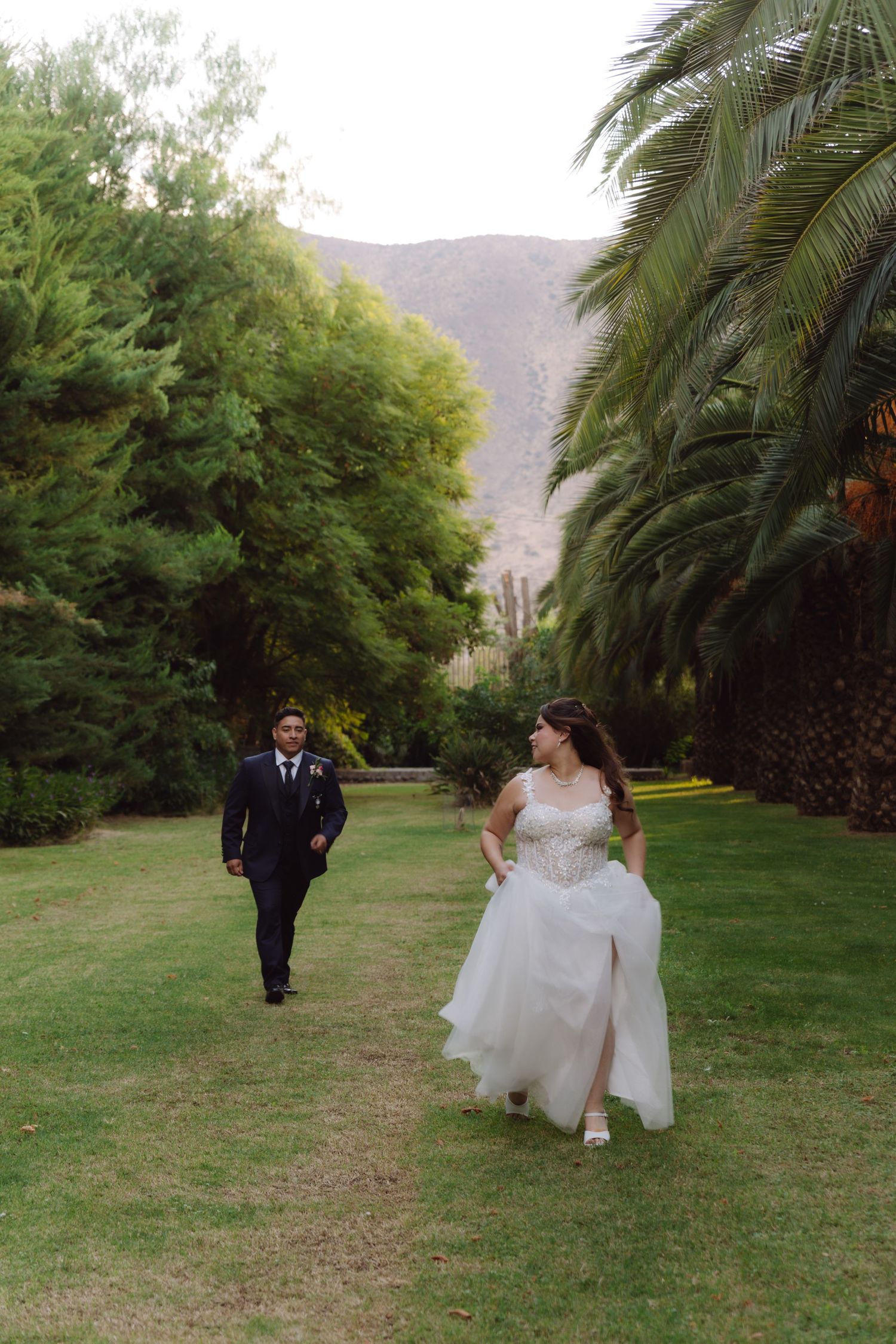 Bride in flowing white gown and groom walking toward each other on a green palm-lined path.