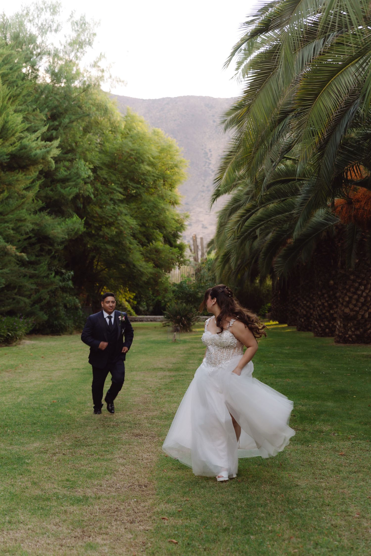 Bride twirling in white dress as groom approaches on a scenic palm tree-lined garden path.