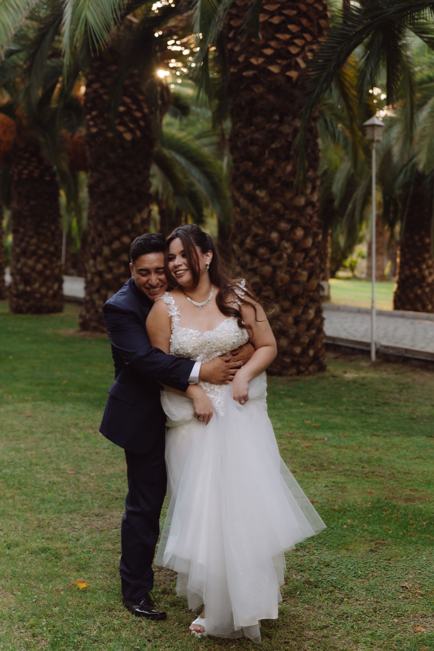 Groom hugging bride from behind as she laughs joyfully in front of tall palm trees.