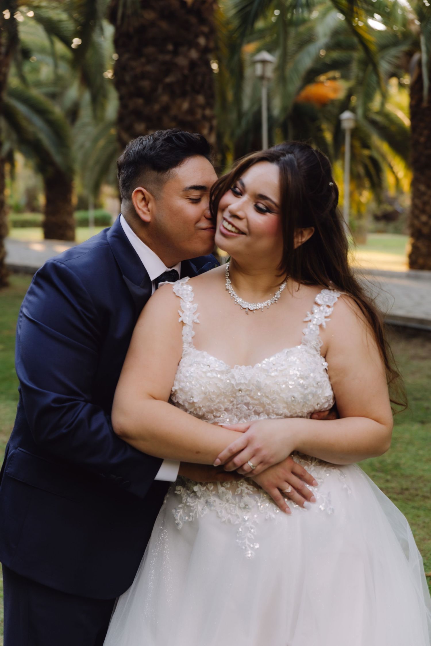 Groom tenderly kissing brides cheek as she smiles warmly in a palm tree garden setting.