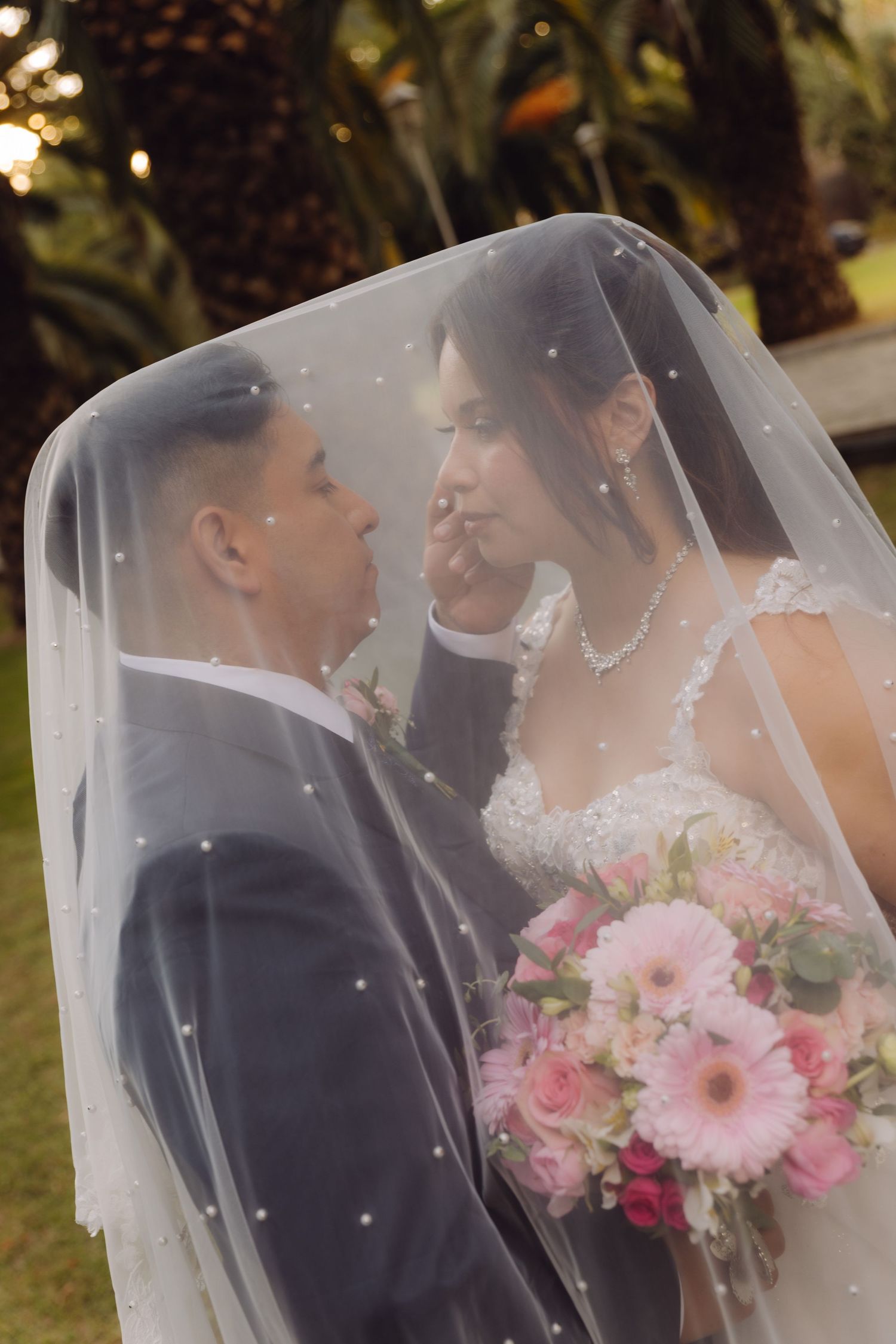 Bride and groom sharing an intimate moment under a dotted veil holding a pink floral bouquet.