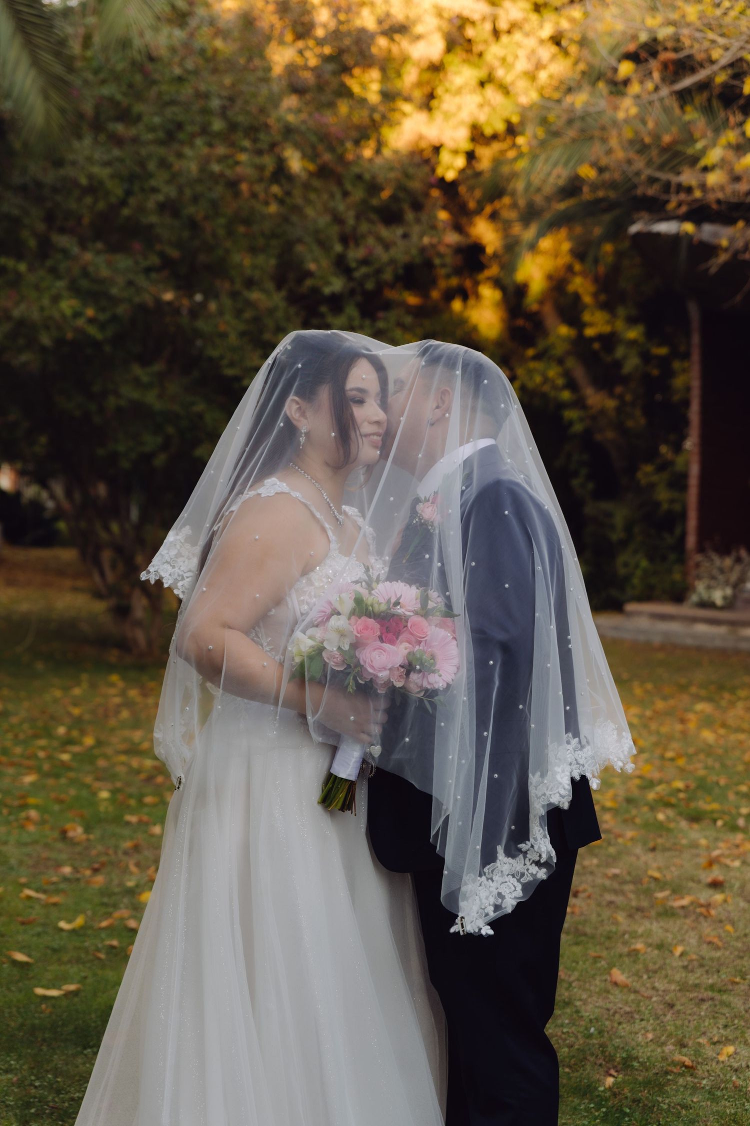 Bride and groom kissing under a lace-trimmed veil in a golden autumn garden setting.