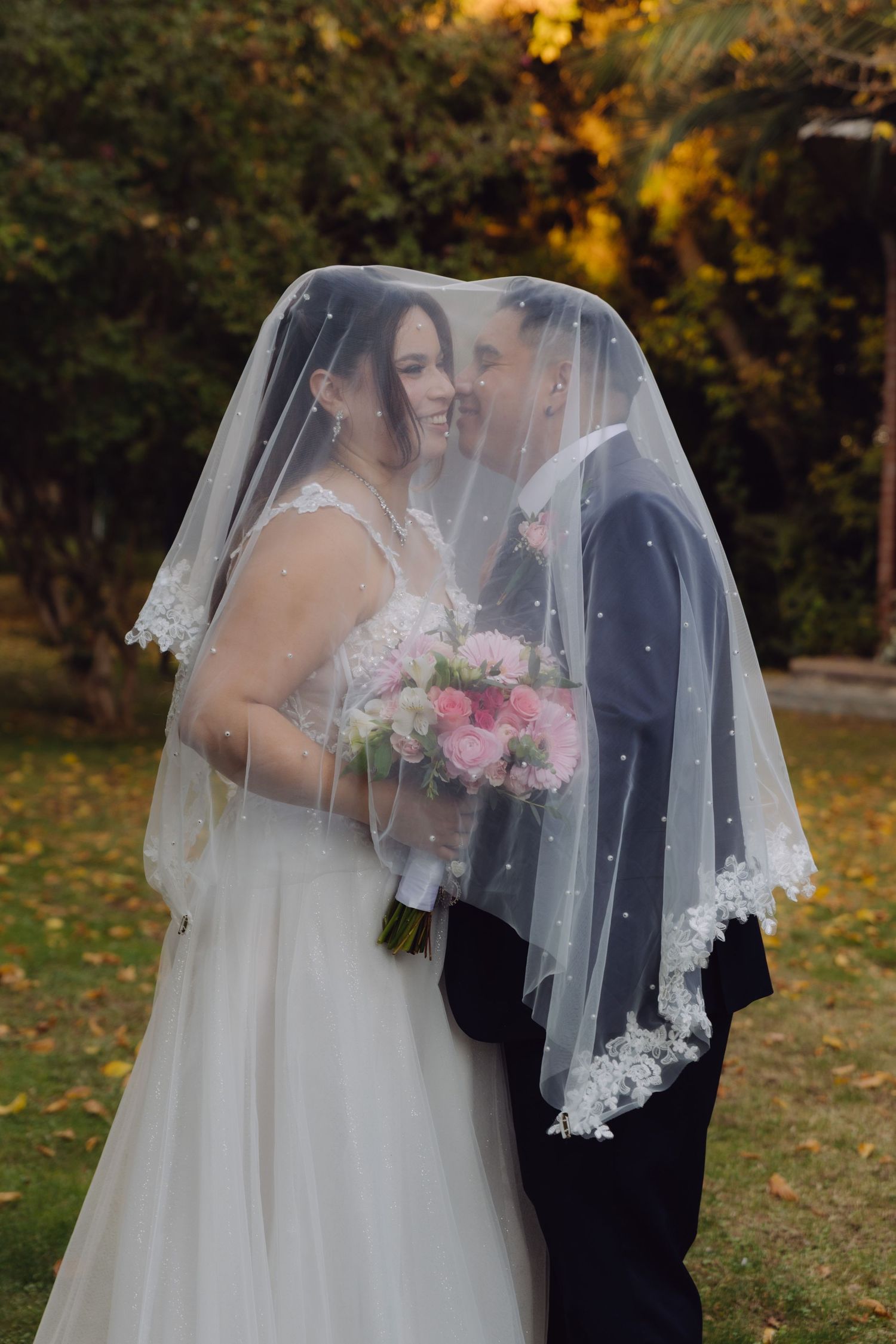 Bride and groom sharing an intimate moment under a pearl-adorned wedding veil outdoors.