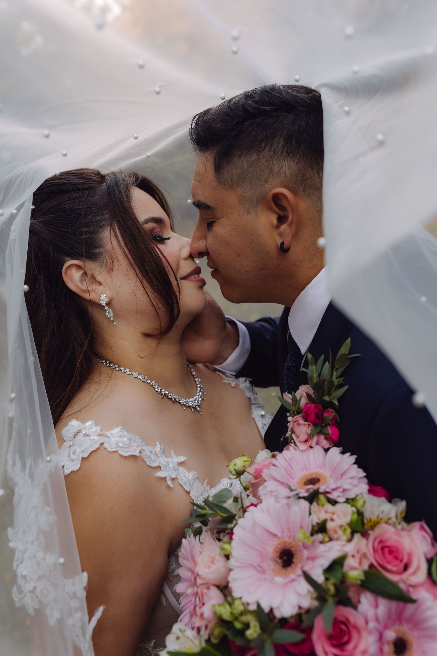 Newlyweds nearly kissing under a pearl veil, bride holding a pink floral bouquet.