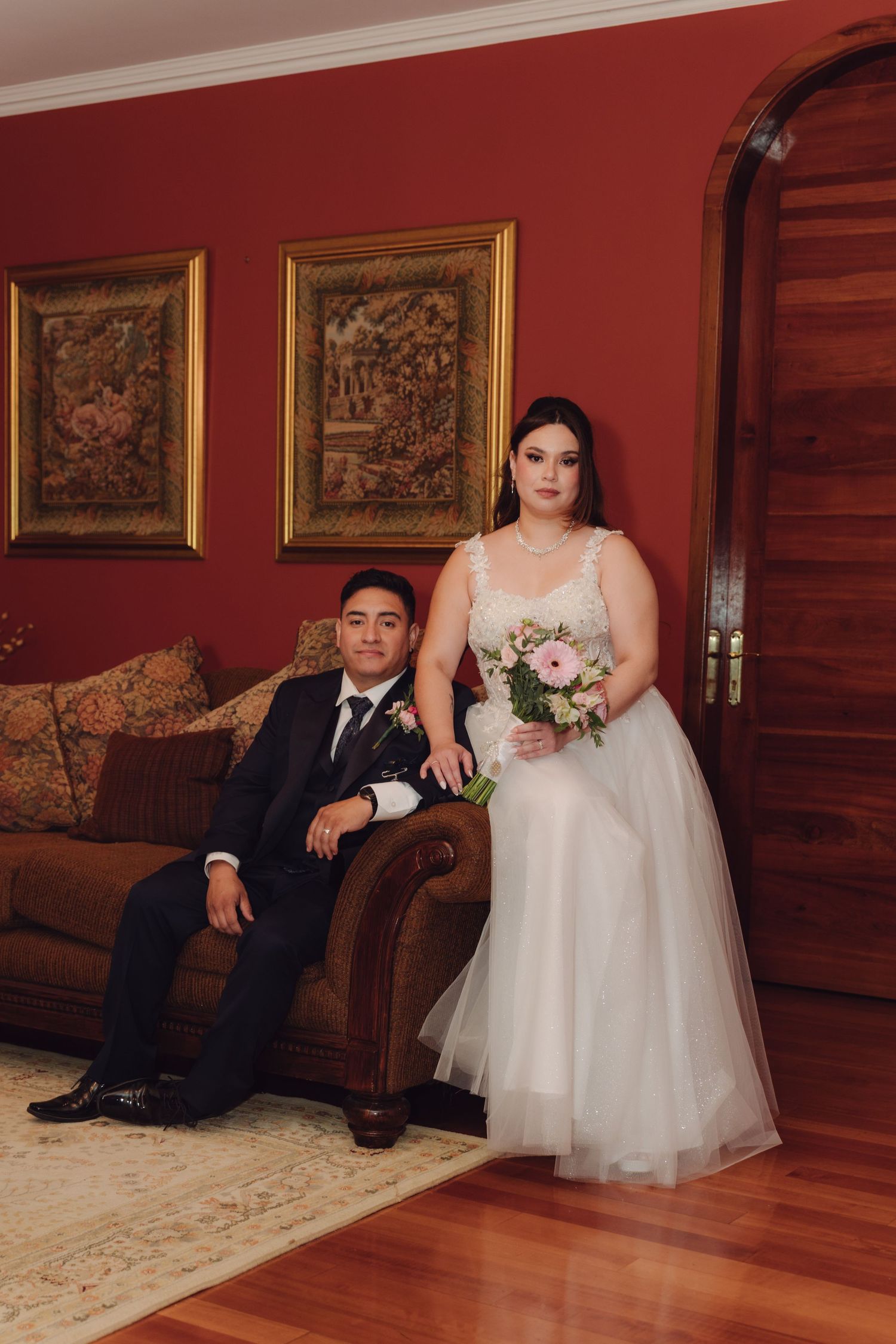 Wedding couple posing indoors, groom seated on sofa, bride standing with pink bouquet.