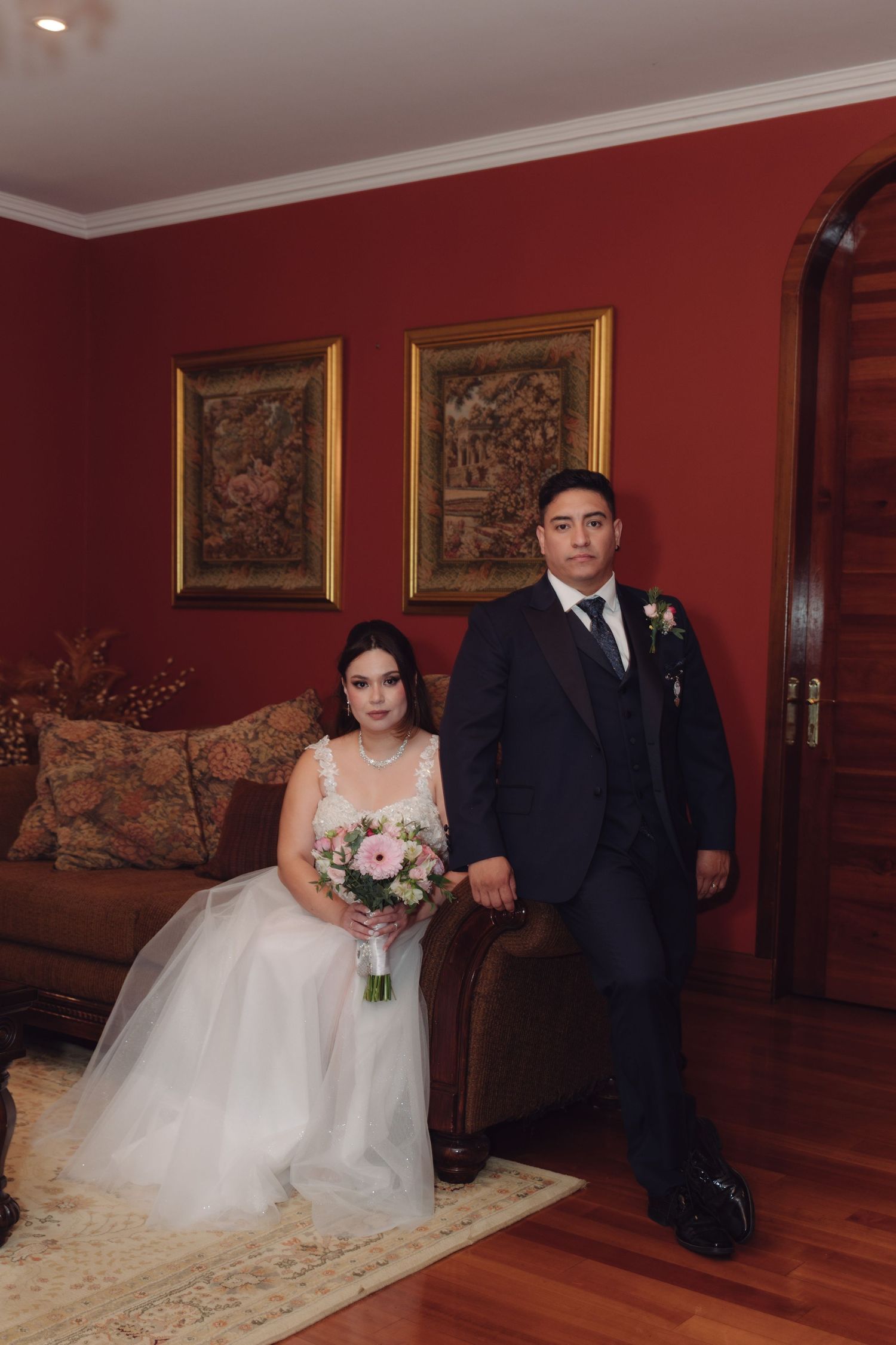 Bride seated on sofa holding bouquet beside groom in navy suit in red-walled room.