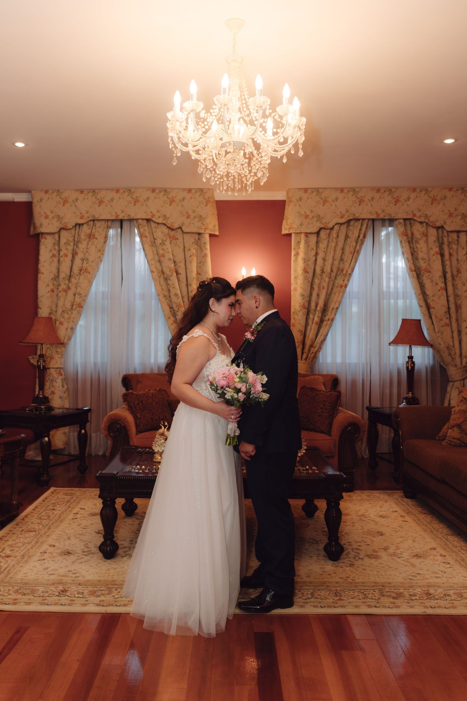 Newlyweds holding pink bouquet facing each other in elegant room under crystal chandelier.
