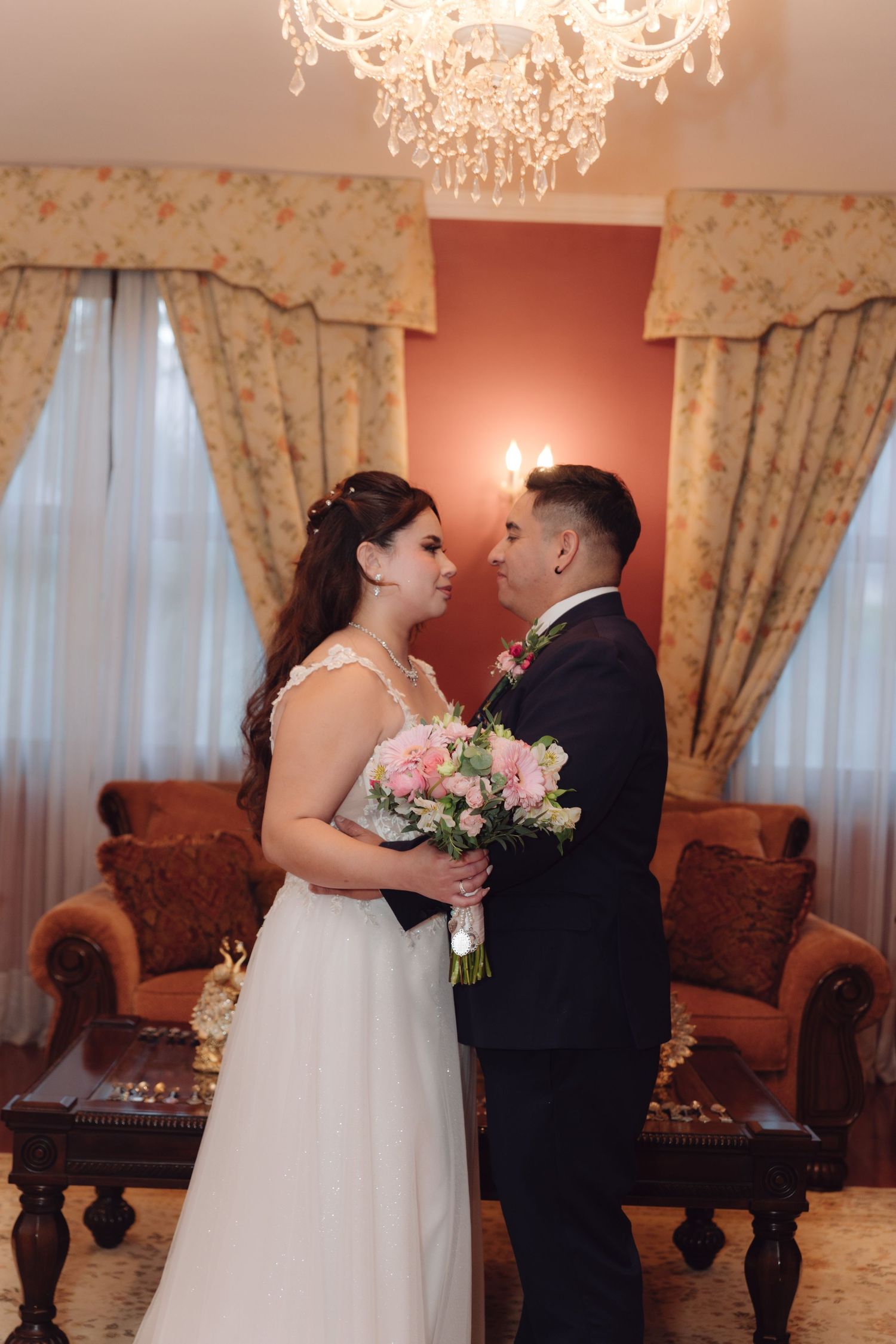 Bride and groom holding floral bouquet gazing at each other under ornate chandelier indoors.