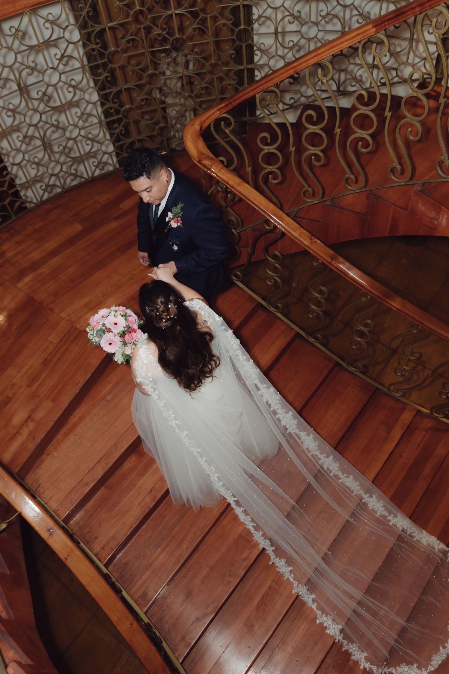 Aerial view of bride and groom holding hands on elegant curved wooden staircase.