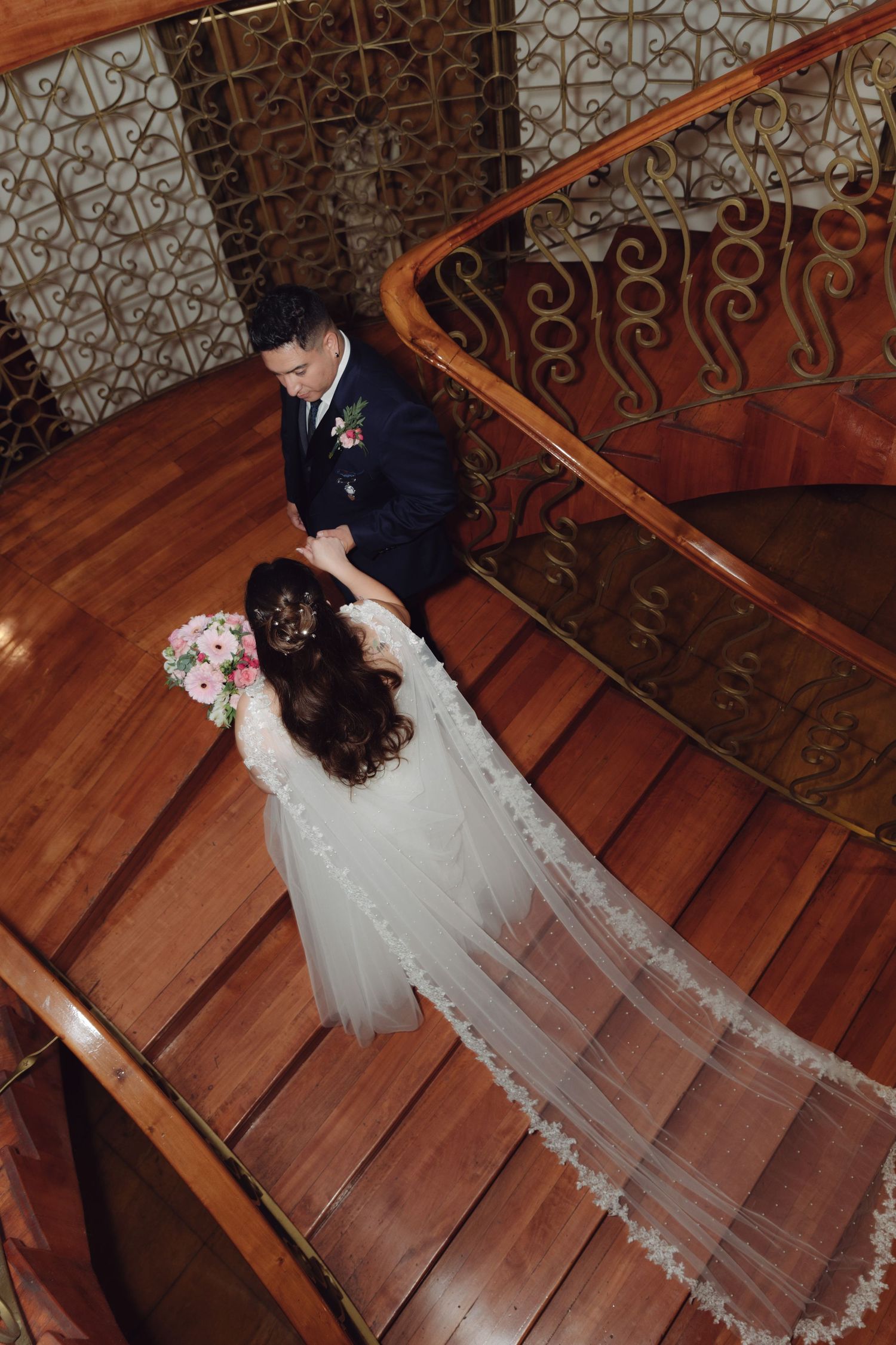 Top-down view of bride with lace veil and groom holding hands on wooden staircase.