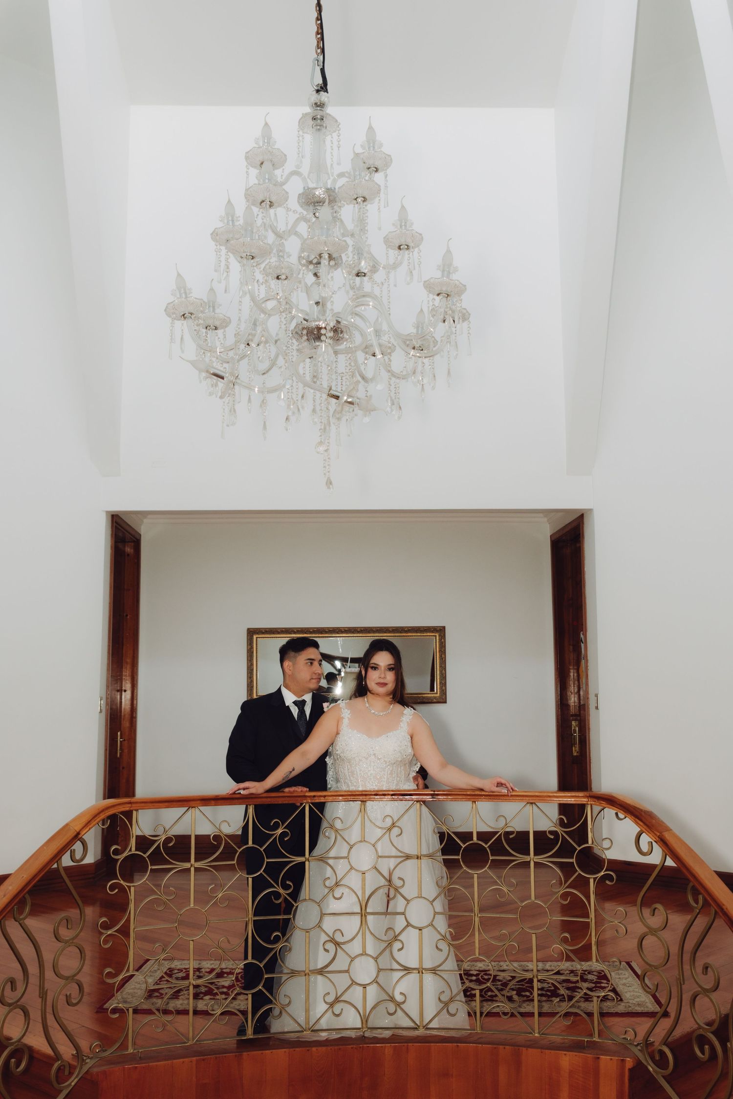Bride and groom pose elegantly on a staircase landing beneath a crystal chandelier in a grand venue.