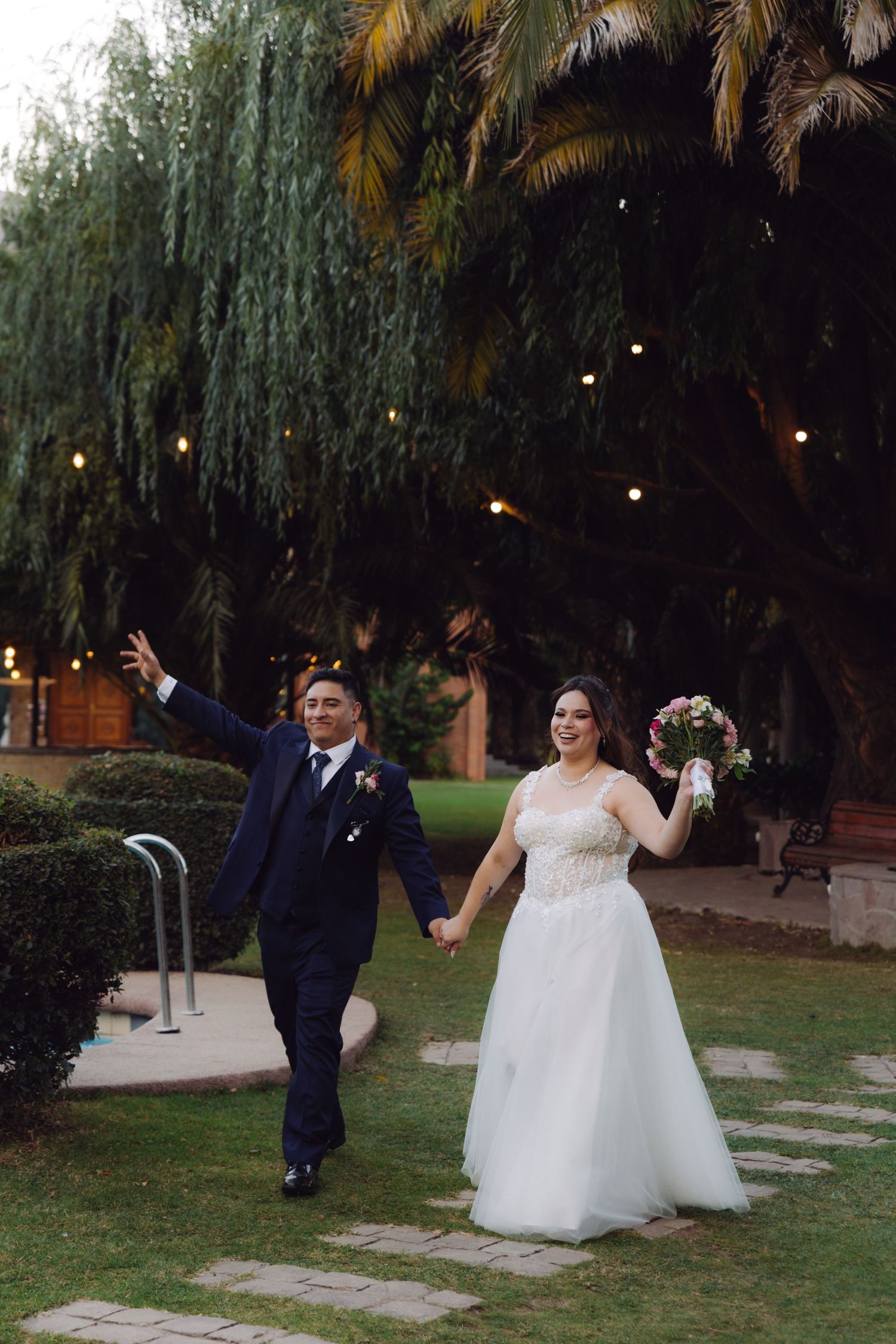 Happy bride and groom walk hand in hand through a garden venue at dusk, bride holding a bouquet.