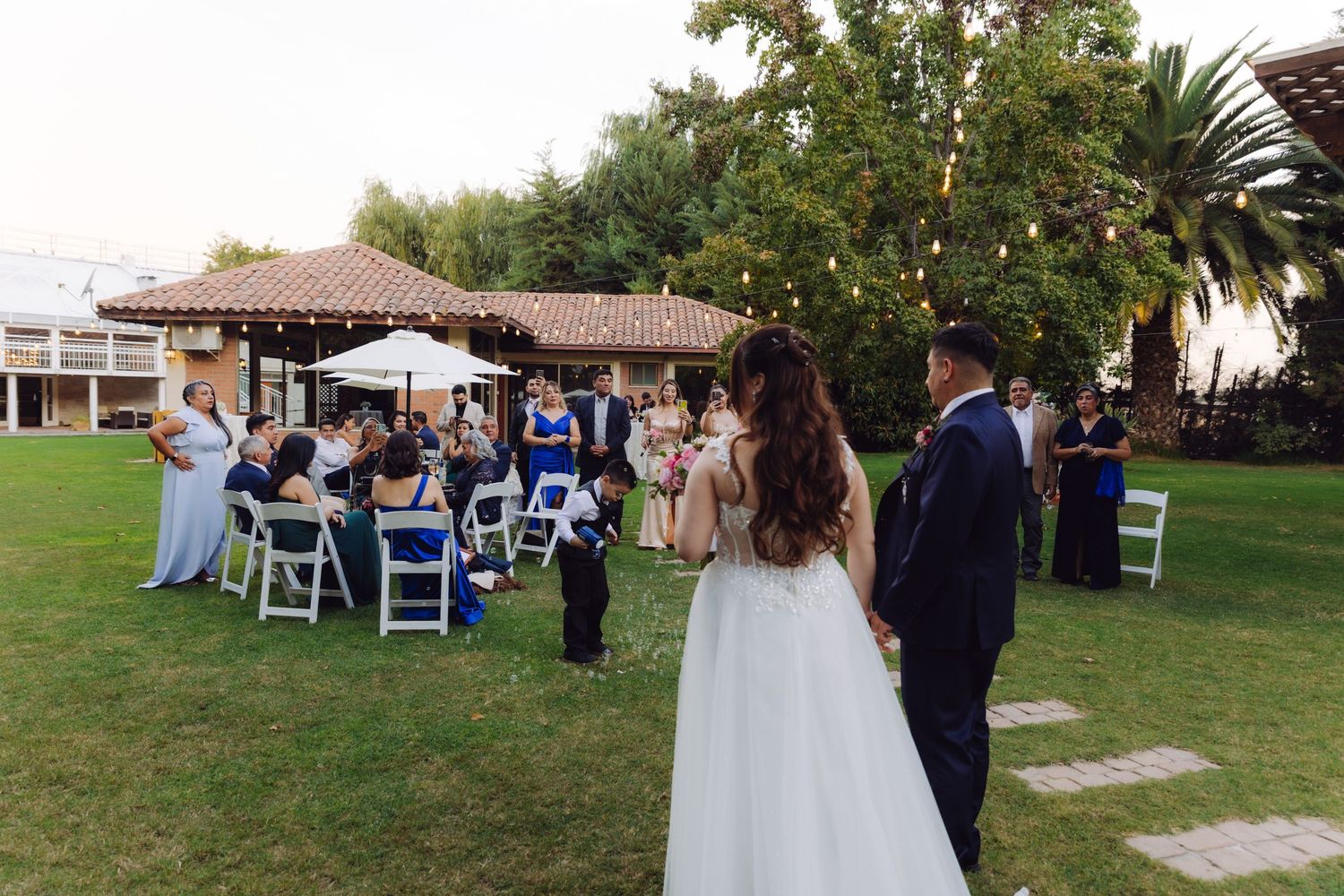 Bride and groom walk toward outdoor wedding guests seated on white chairs in a garden ceremony.