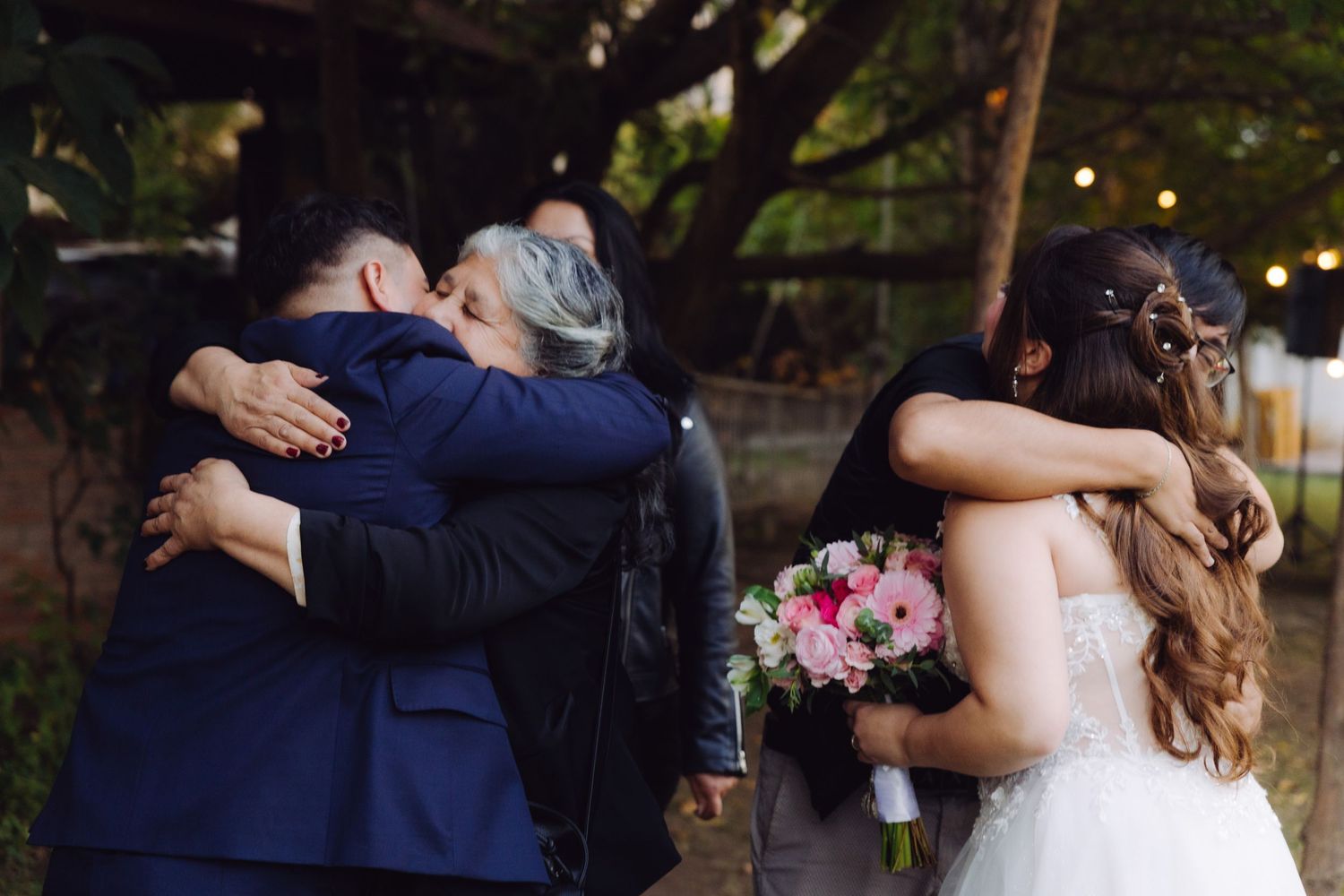 Bride and groom embrace family members in an emotional outdoor wedding reception moment.