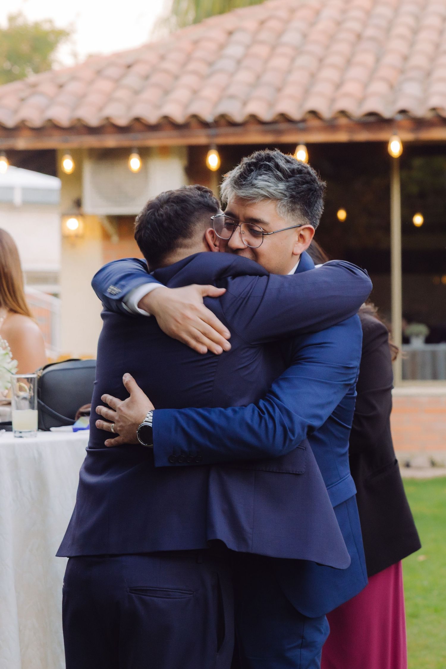 Two suited men share a warm emotional embrace at an outdoor wedding reception celebration.