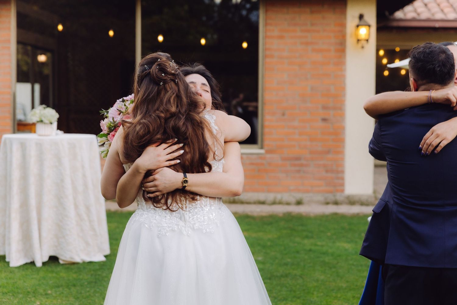 Bride joyfully hugs a female guest while groom embraces another guest at outdoor reception.
