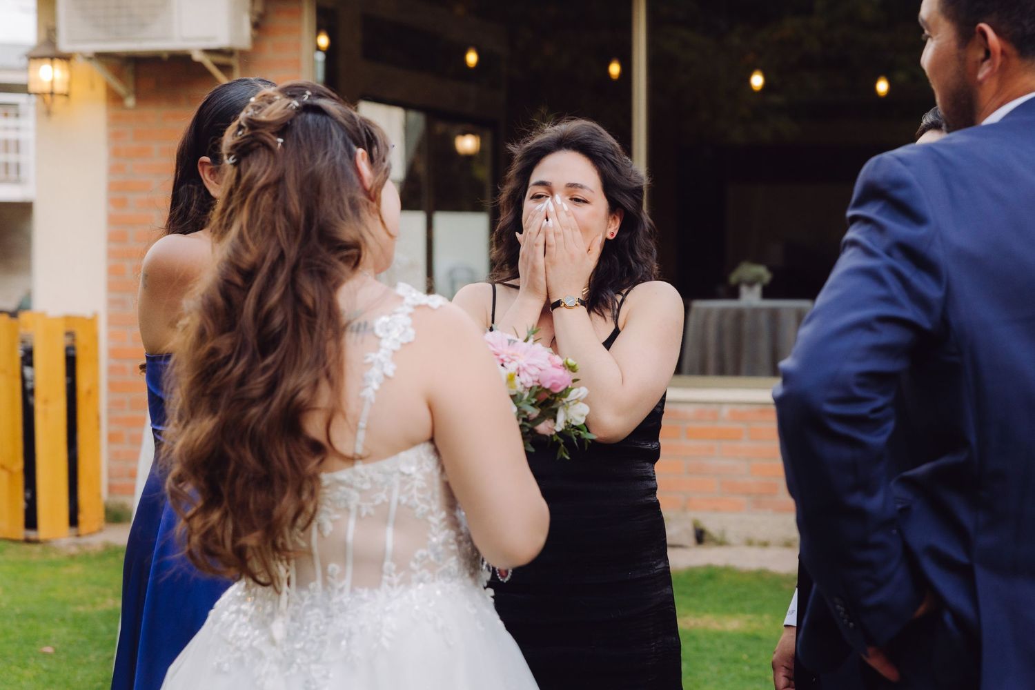 Emotional wedding guest covers mouth in surprise while greeting bride at outdoor reception.