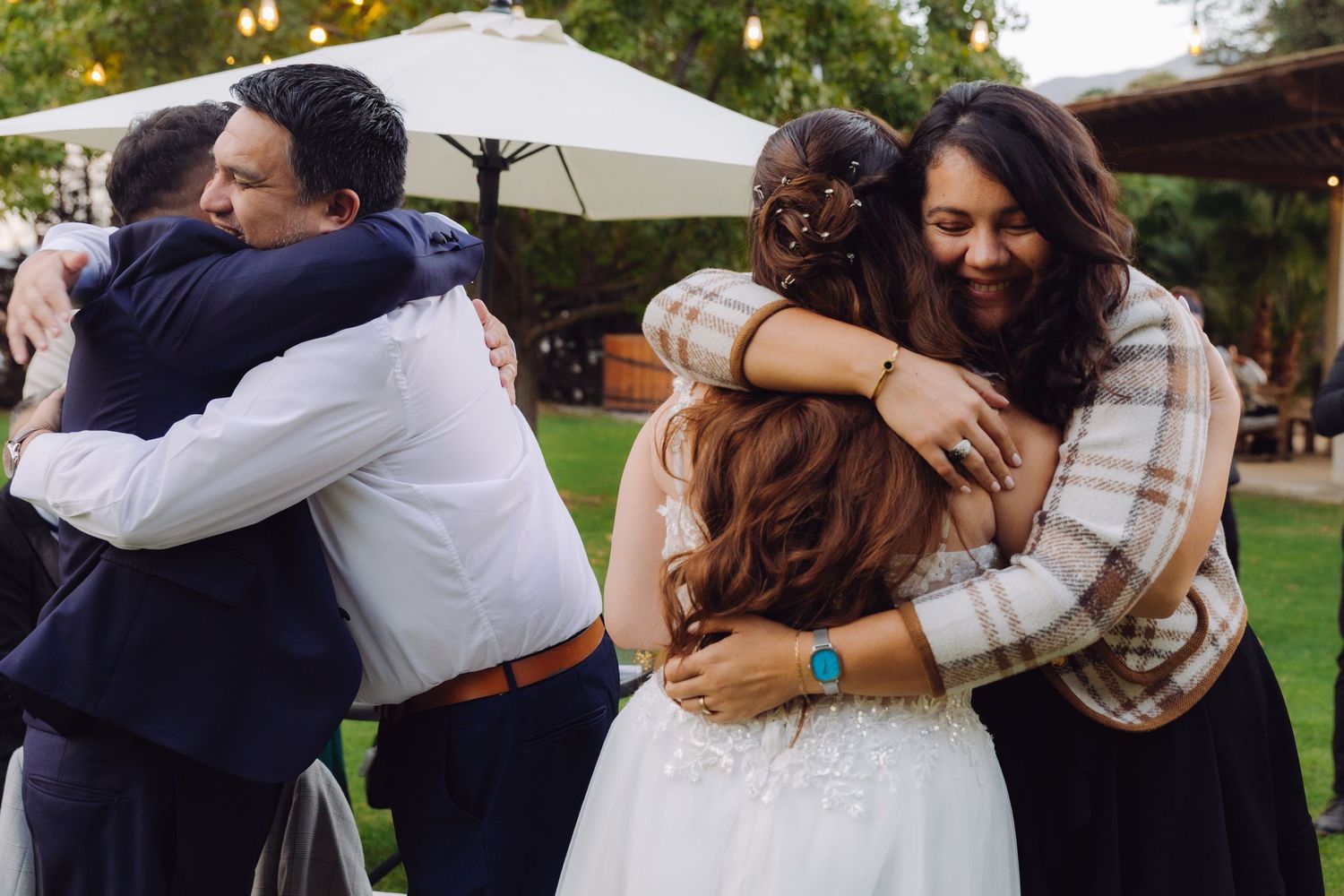 Joyful group hug between bride, groom, and wedding guests on lawn after ceremony.
