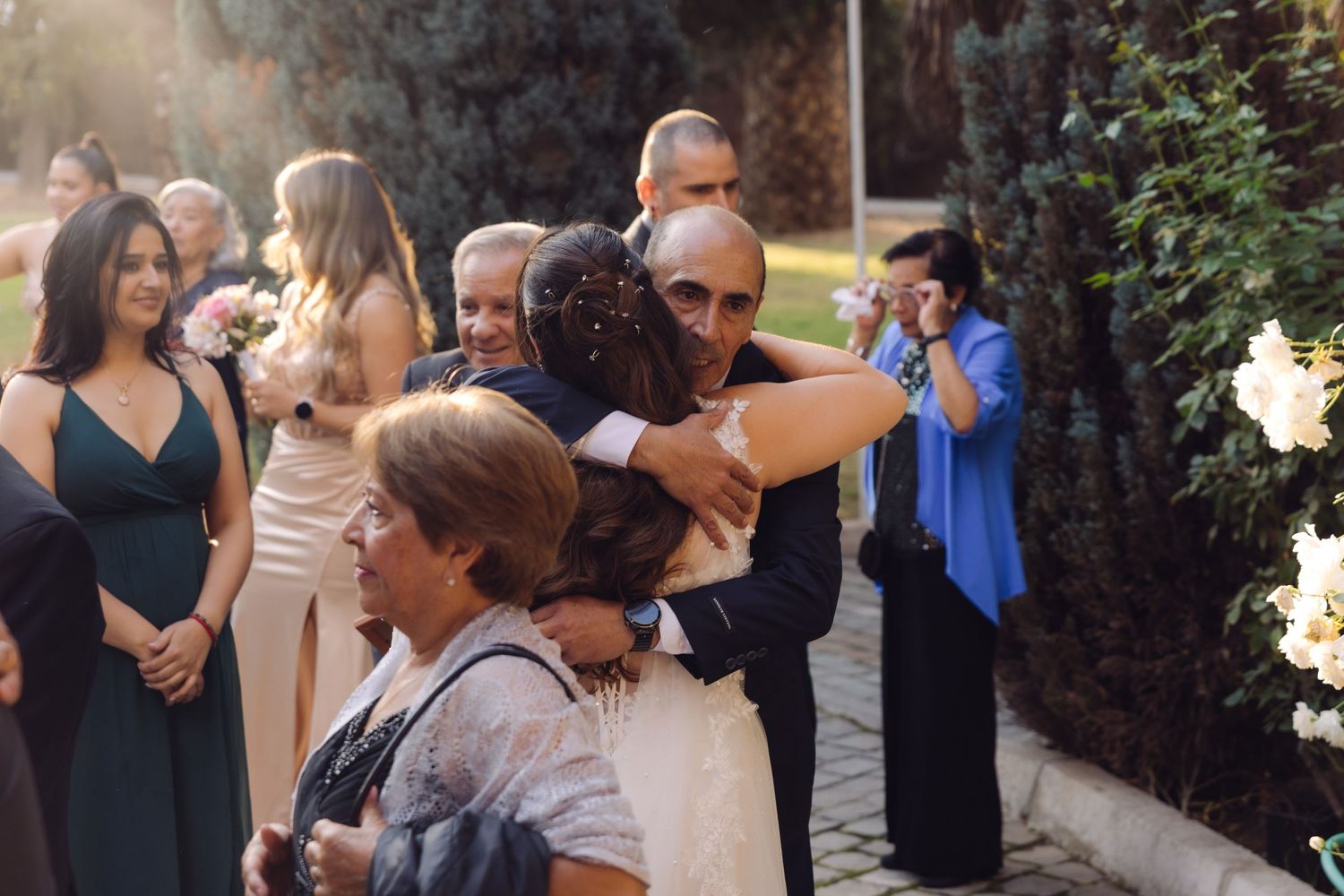 Bride receives warm embrace from bald guest while wedding party mingles in garden setting.