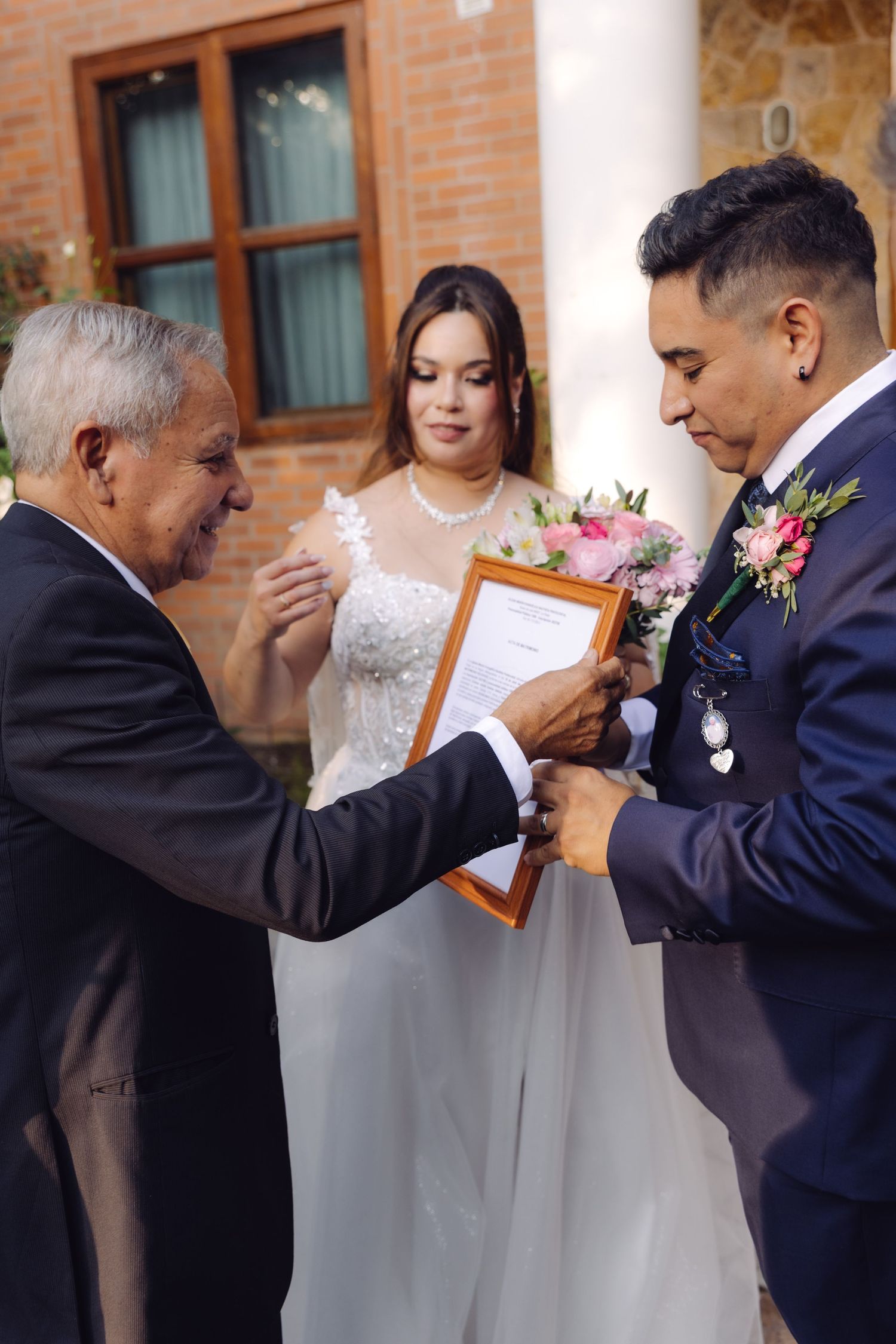 Elder gentleman presents framed wedding certificate to groom as bride watches at ceremony.