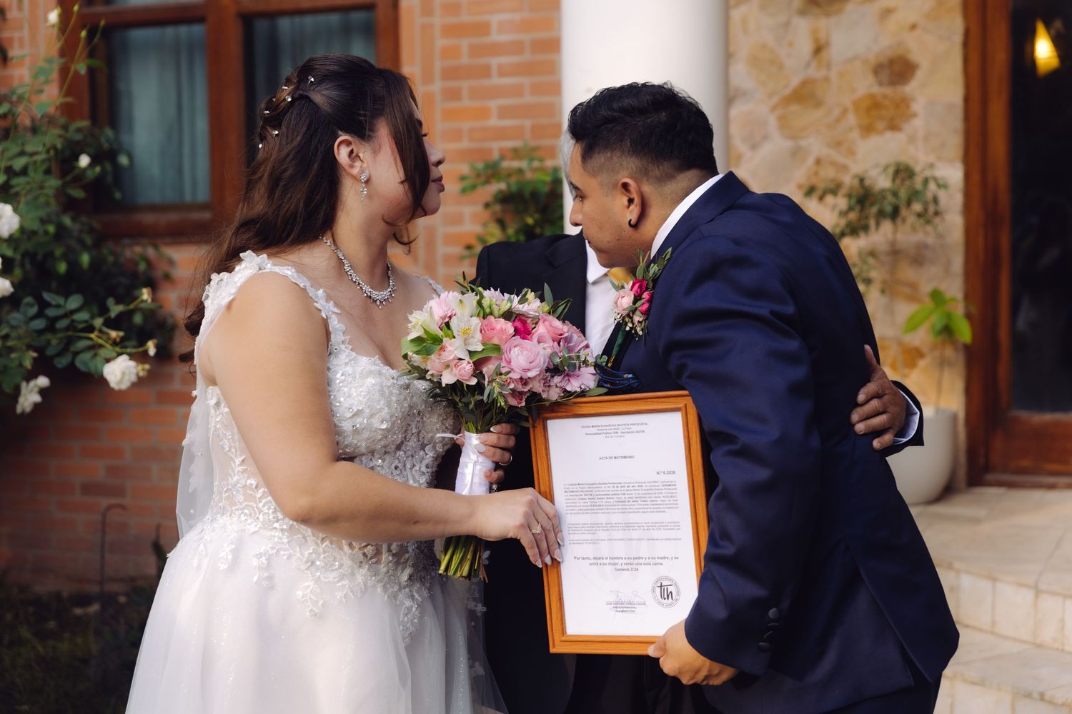 Bride and groom share tender hug while groom holds framed wedding certificate outdoors.