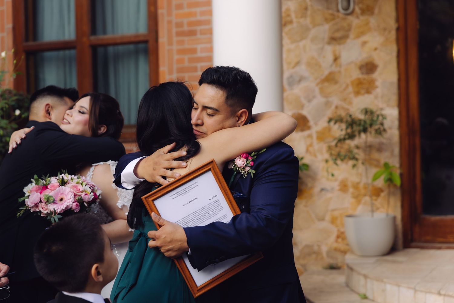 Groom embraces guest while holding framed wedding certificate outside rustic stone venue.