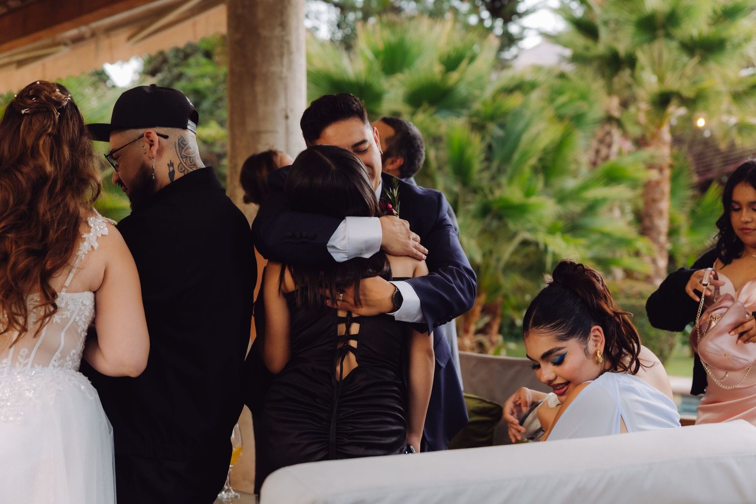 Groom shares heartfelt hug with guest under outdoor pavilion surrounded by wedding party.
