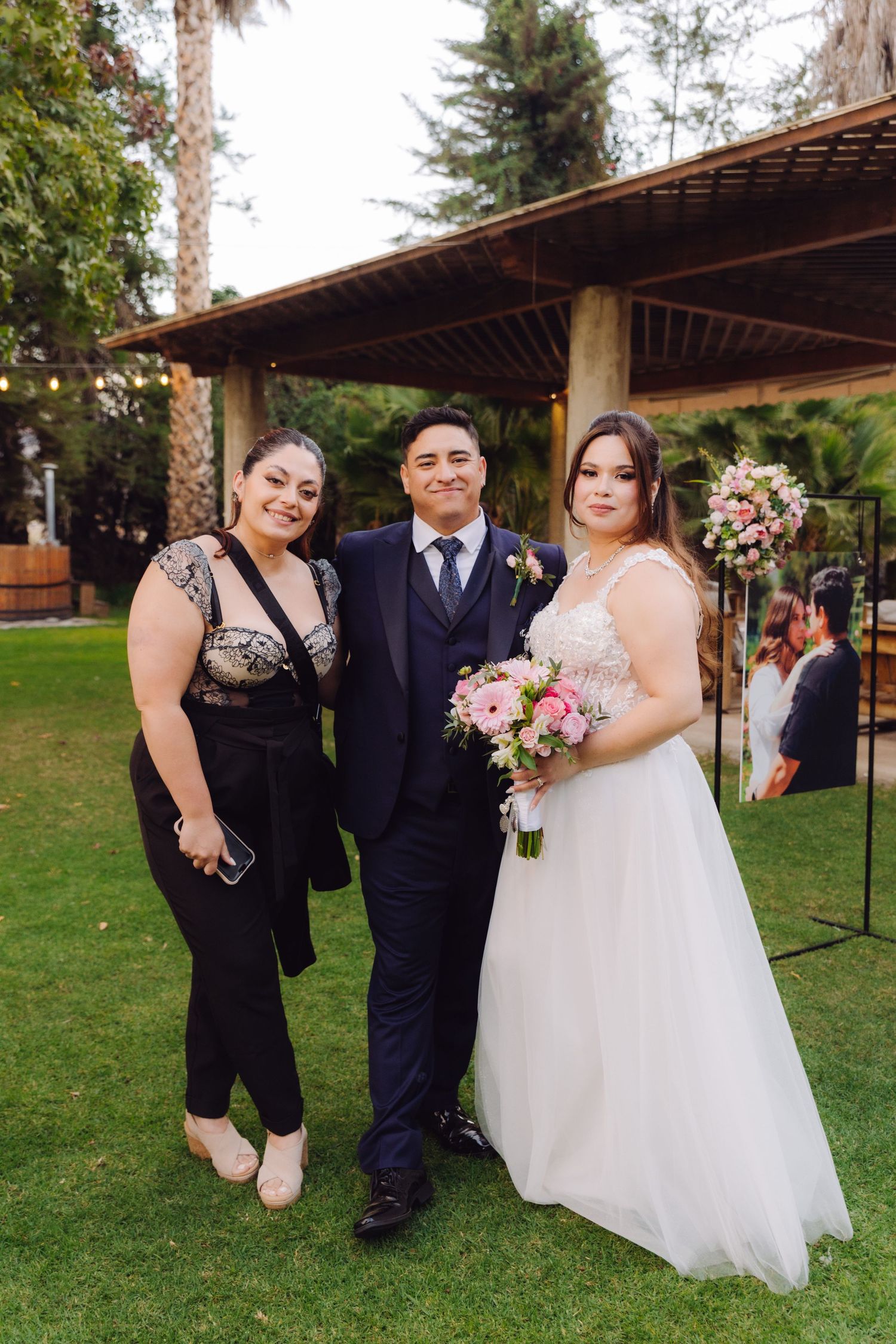 Bride in white gown holds pink bouquet posing with groom in navy suit and woman in black at outdoor wedding.