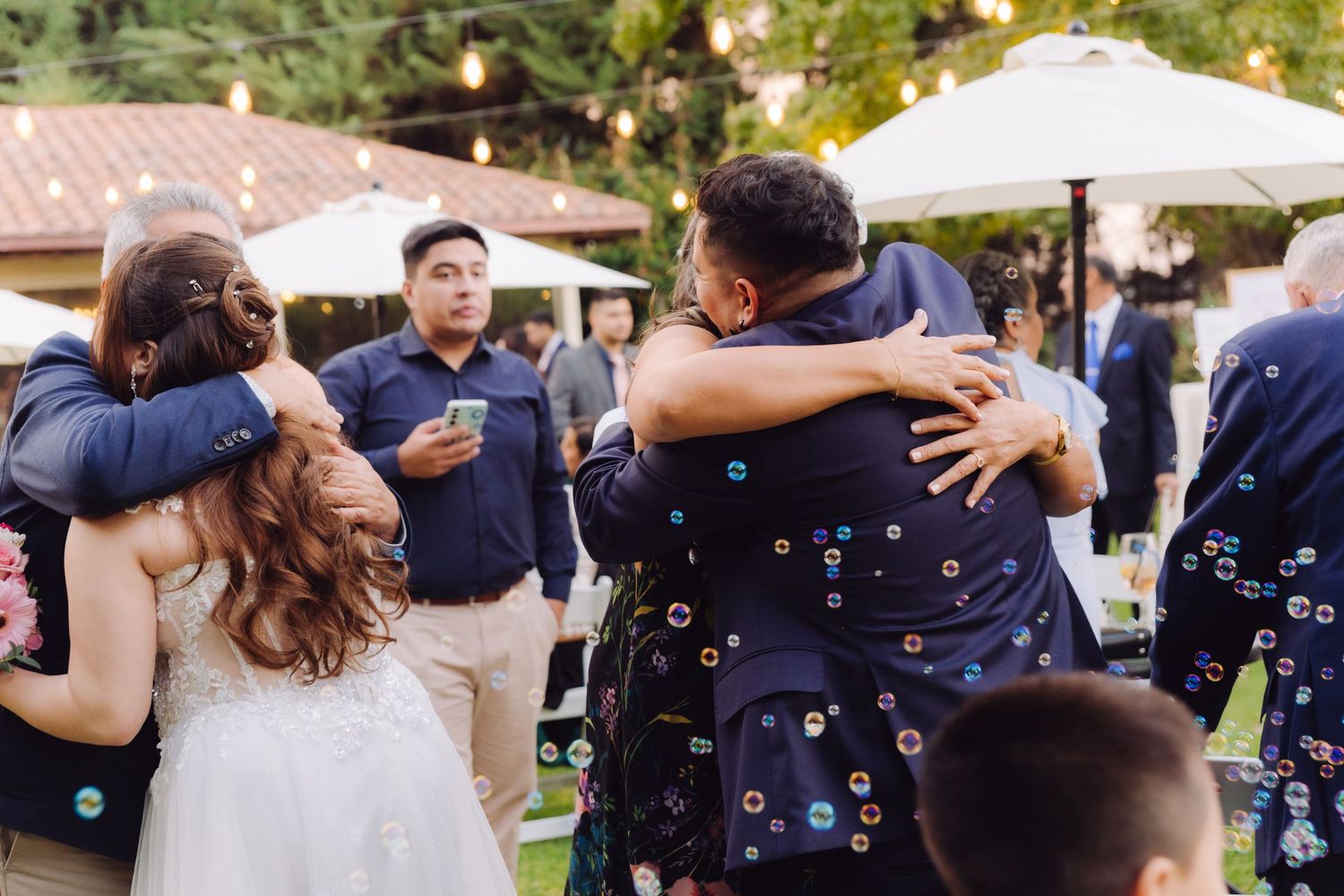 Wedding guests share warm embraces outdoors with string lights and white umbrellas in background.
