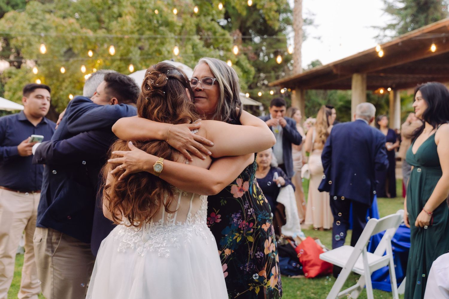 Bride in lace wedding gown receives heartfelt hug from older woman guest at outdoor wedding reception.