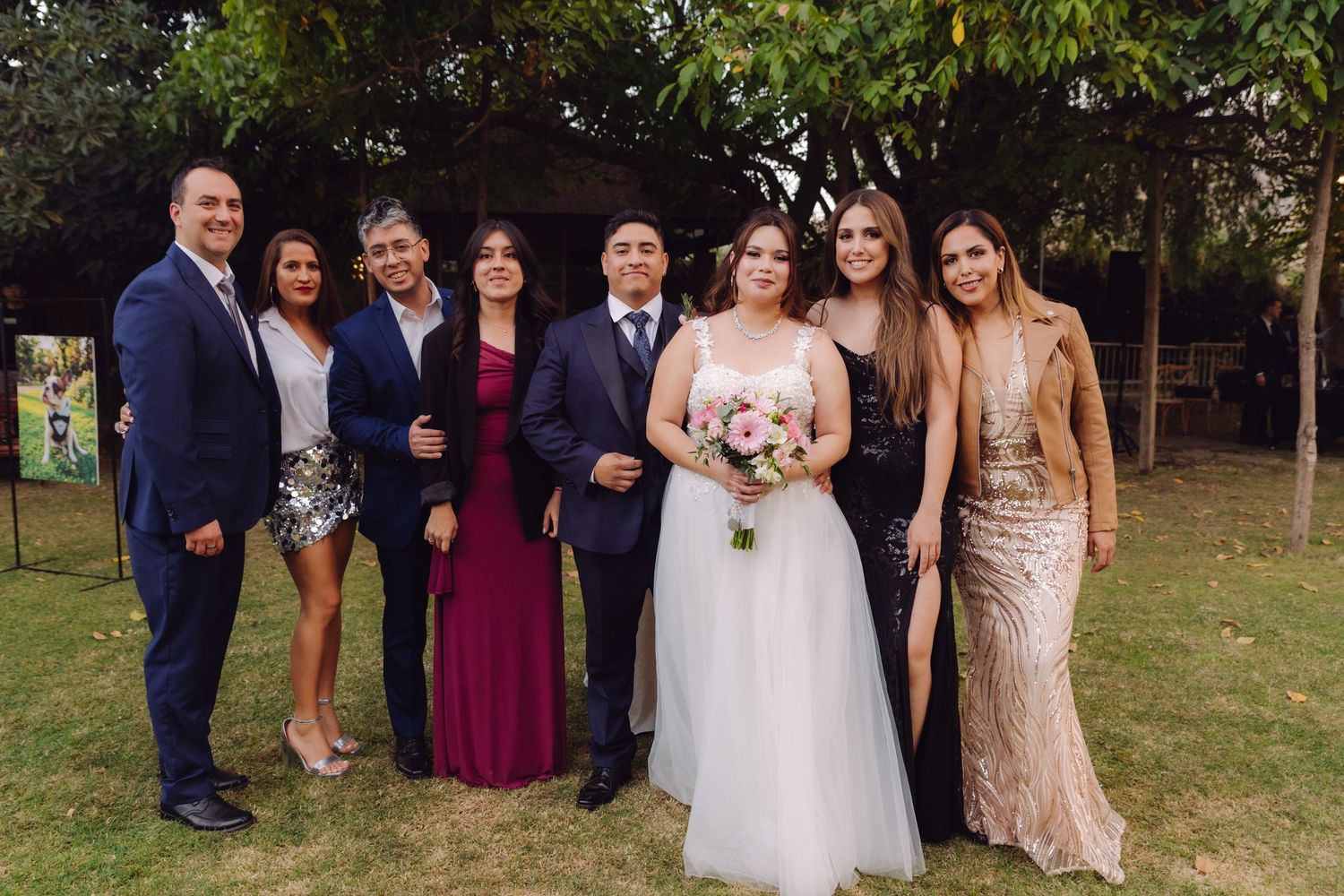 Wedding party group portrait on green lawn with bride holding pink bouquet surrounded by elegantly dressed guests.
