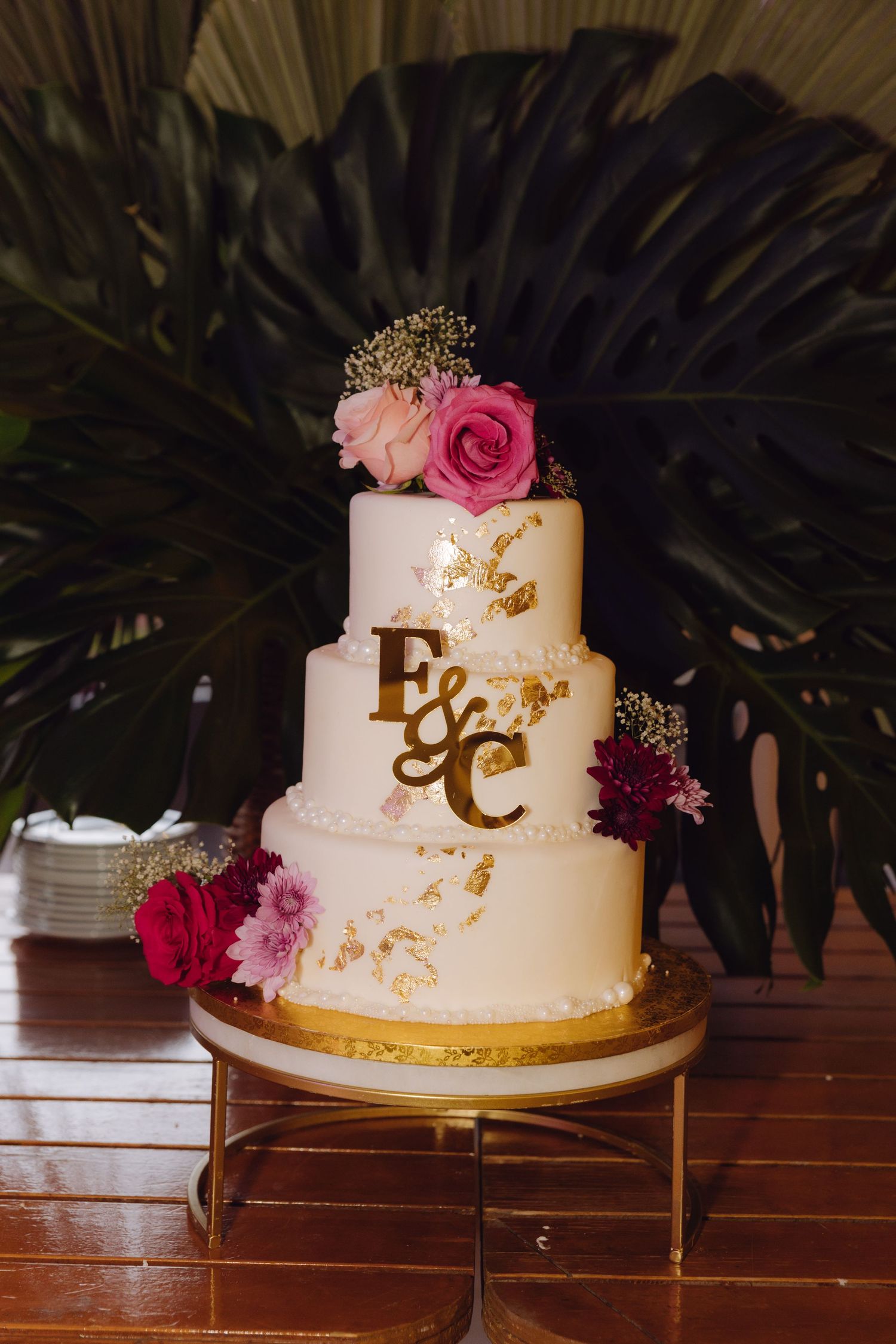 Three-tier white wedding cake with gold E&C monogram decorated with pink and burgundy flowers on gold stand.