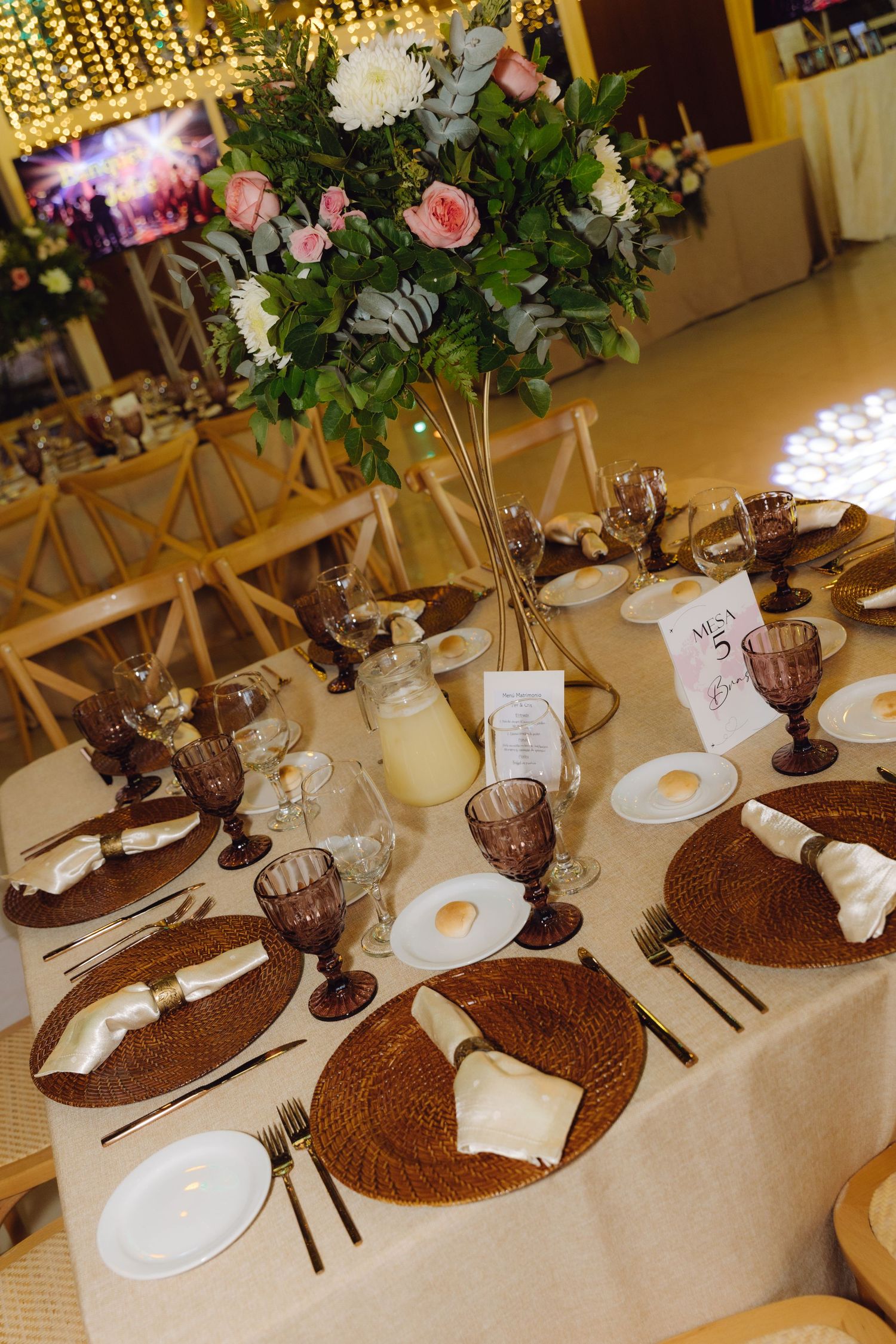 Elegant wedding reception table set with woven chargers, purple goblets, white plates, and tall floral centerpiece.