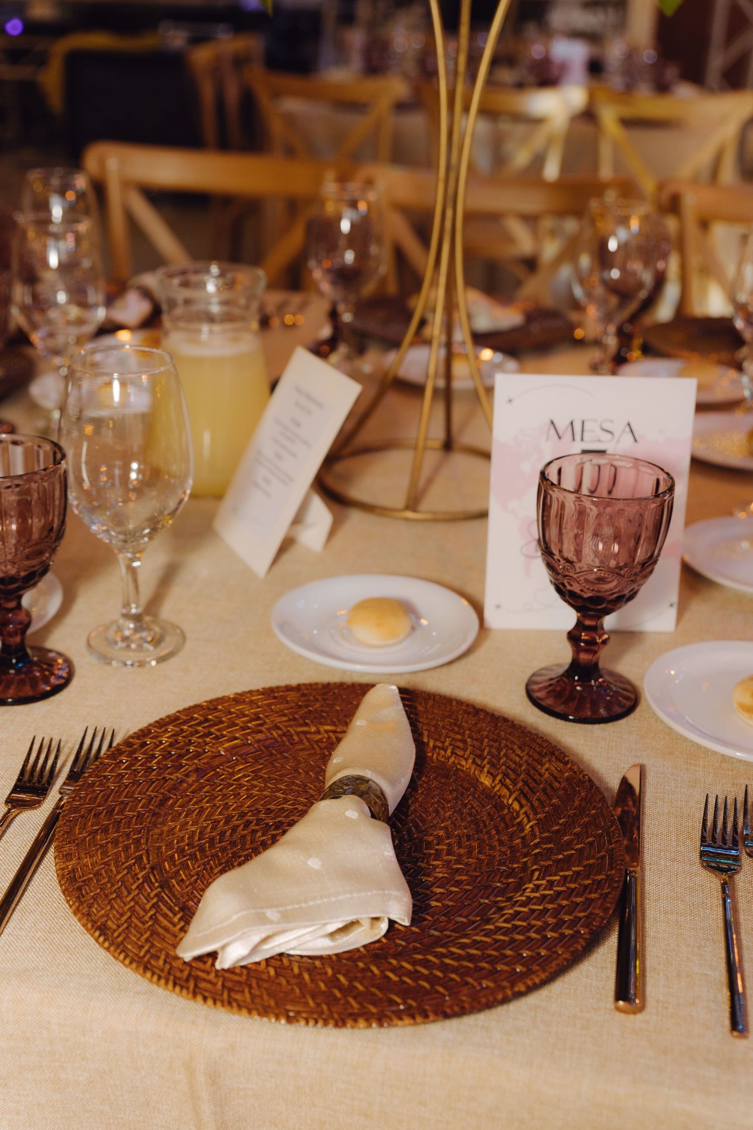Close-up of wedding table place setting with woven charger plate, folded napkin, purple goblet, and Mesa card.
